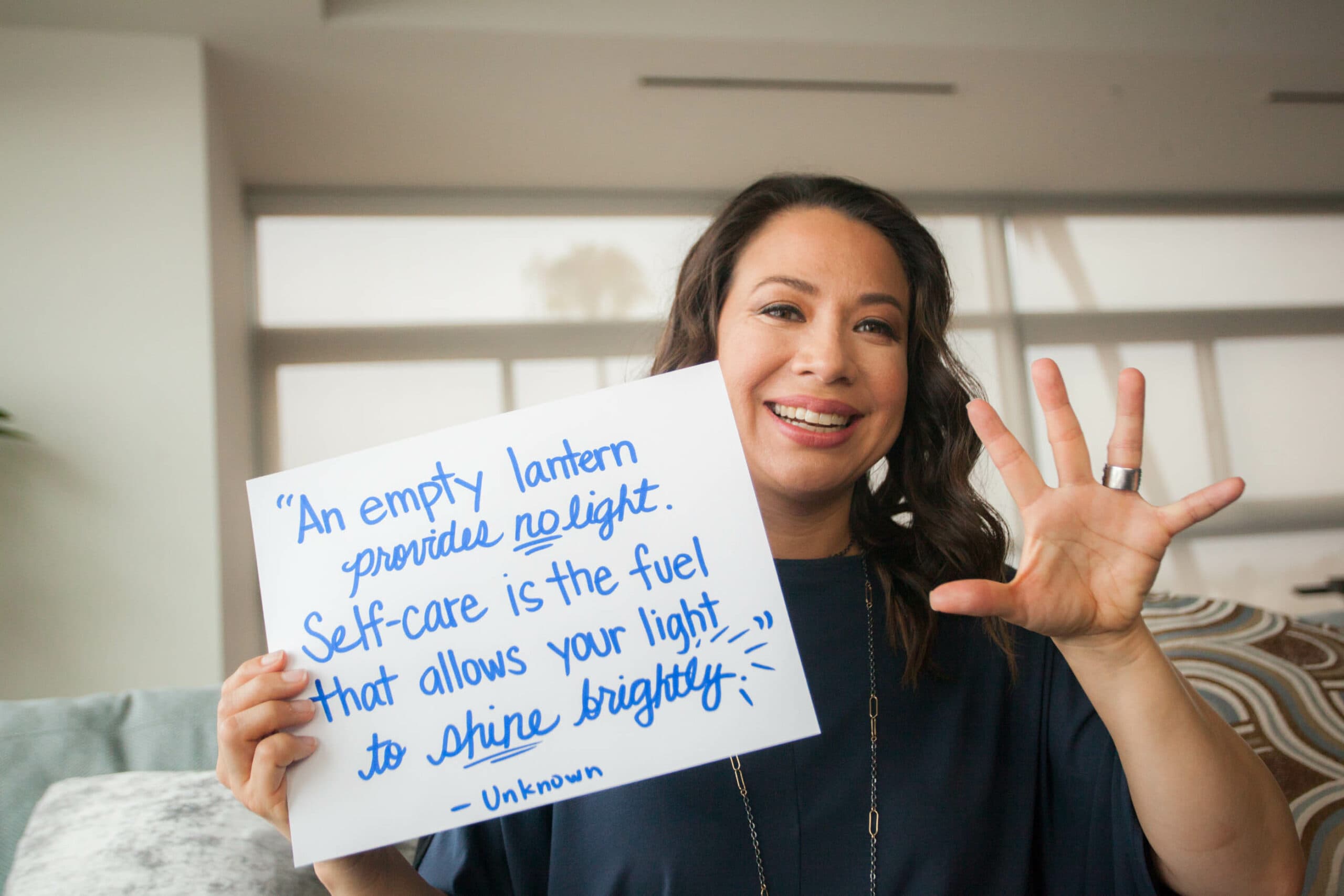 A woman smiling and holding up a handwritten sign that says, An empty lantern provides no light. Self-care is the fuel that allows your light to shine brightly. She is gesturing with her other hand, showing four fingers.