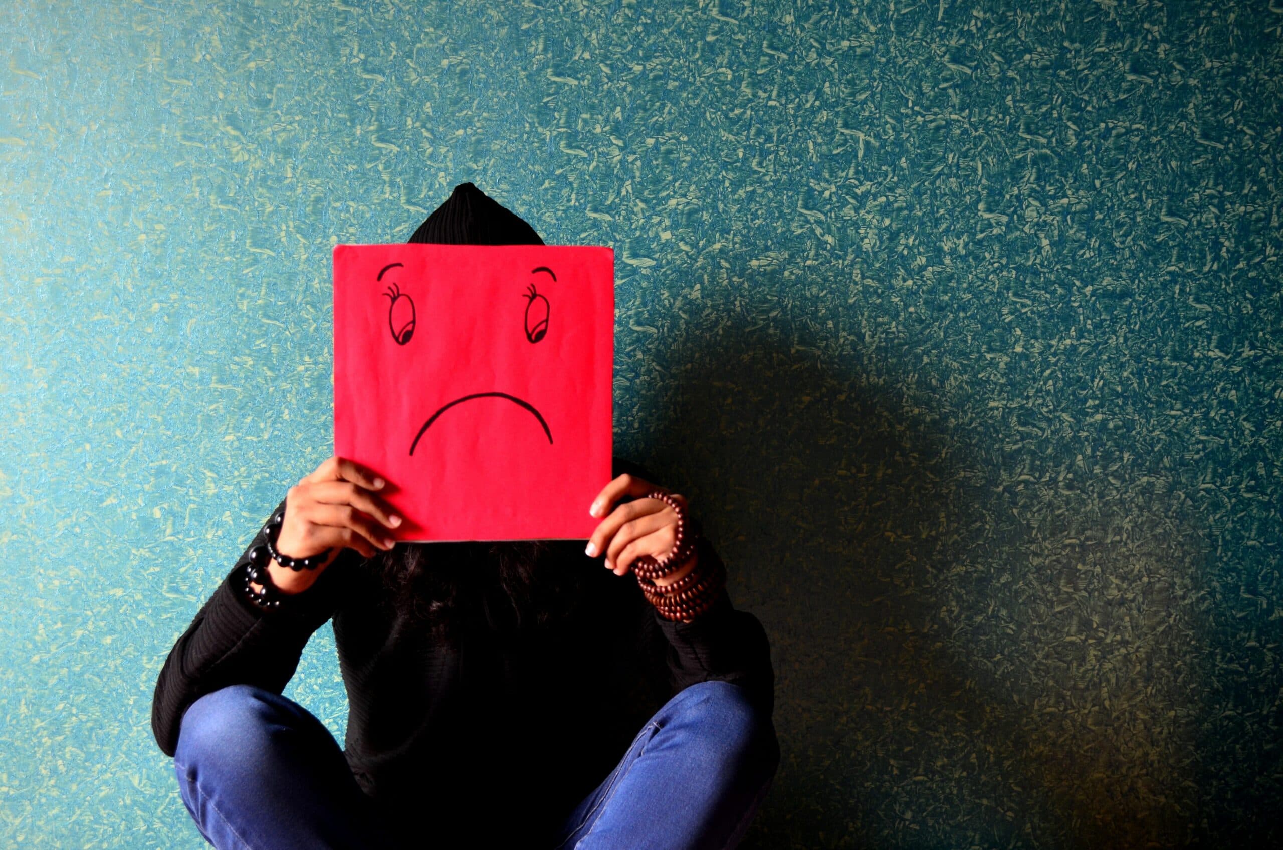 A person sitting against a textured teal wall, holding a red square with a drawn sad face in front of their head. They wear a black hoodie and blue jeans.