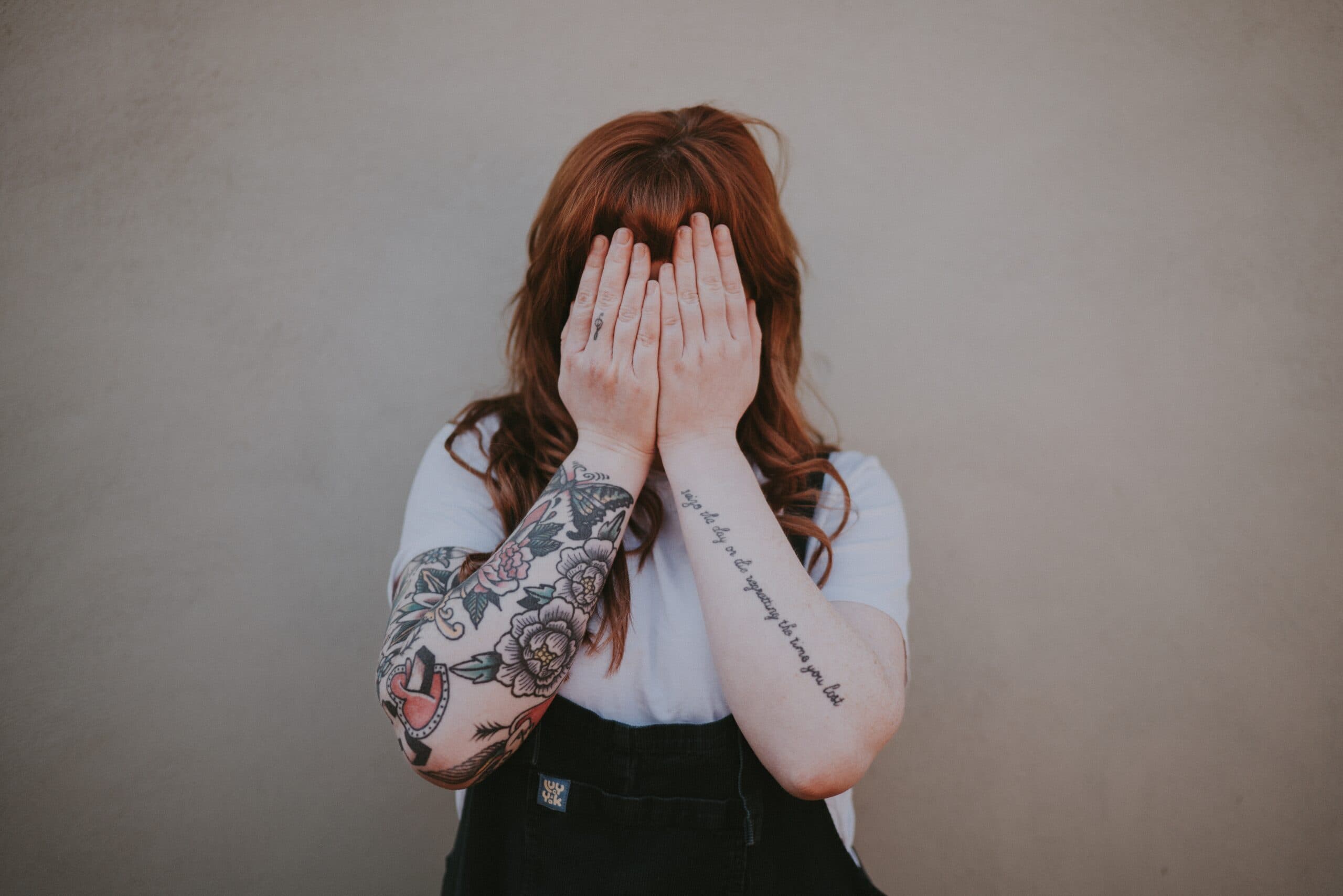 A person with long red hair covers their face with both hands. They are wearing a white shirt and black overalls. Visible on their arms are tattoos featuring flowers and text, standing against a neutral beige background.