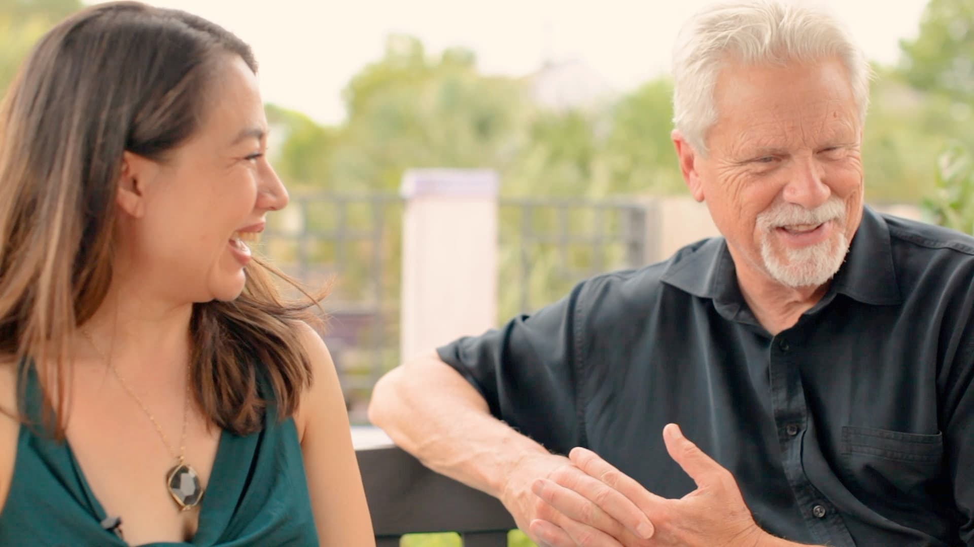 A woman and an older man are sitting together on a bench, laughing and smiling at each other. The woman has long brown hair and is wearing a green sleeveless top. The man has white hair, a beard, and is wearing a black shirt. Background is blurred greenery.