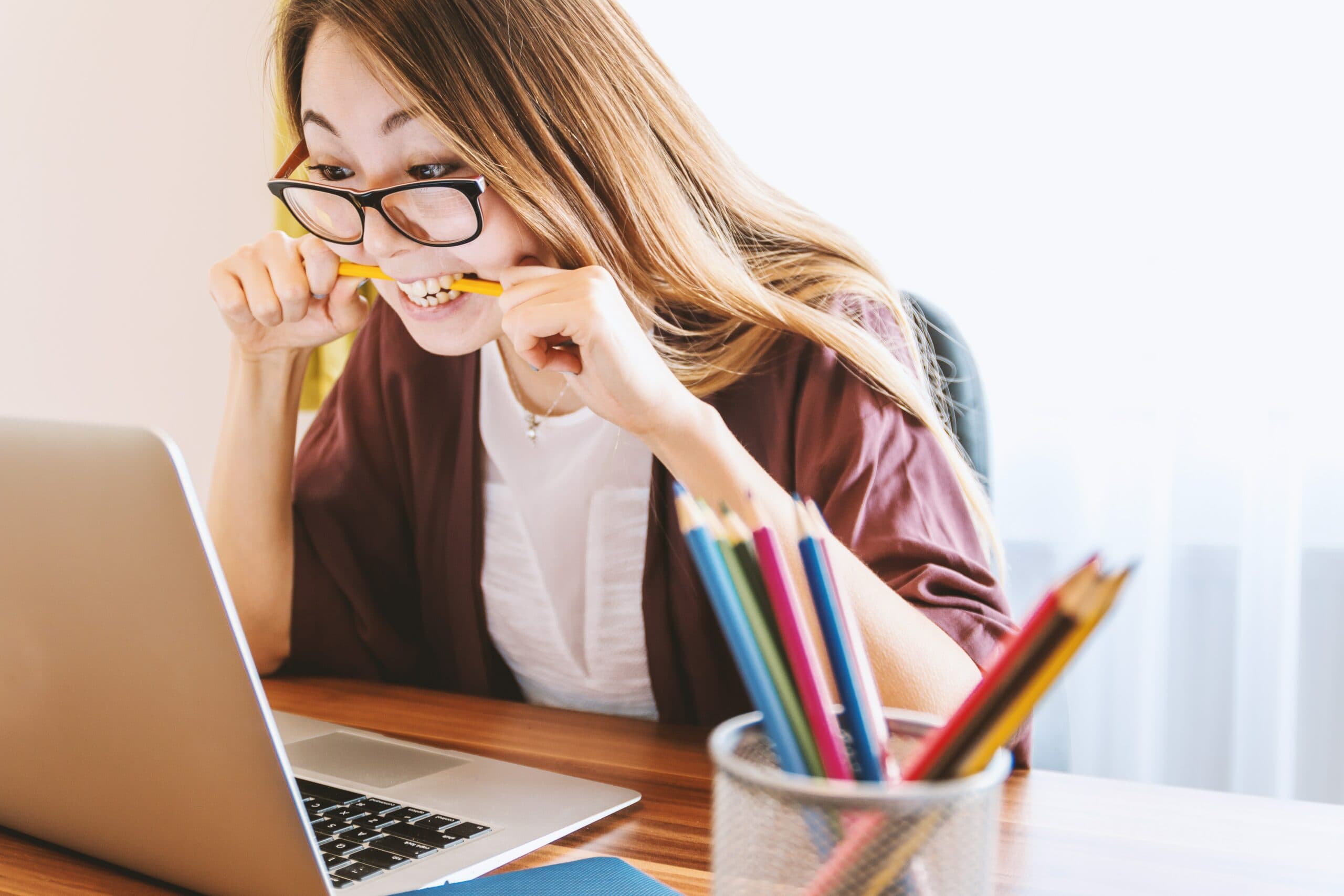 A woman with long hair and glasses is sitting at a desk, biting a pencil while looking at a laptop. A cup filled with colored pencils is on the desk in front of her, and natural light comes through a window in the background.