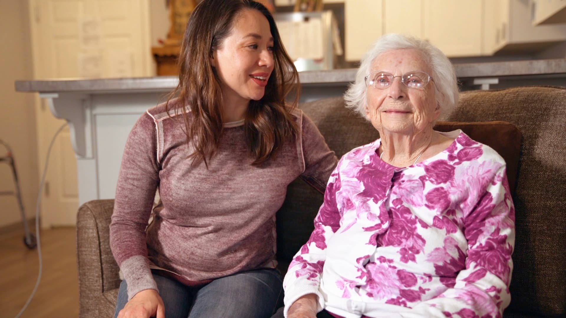 A young woman converses with an elderly woman seated on a couch. The young woman wears a brown sweater and jeans, and the elderly woman wears glasses and a floral pink and white top. They are in a cozy room with a kitchen in the background.