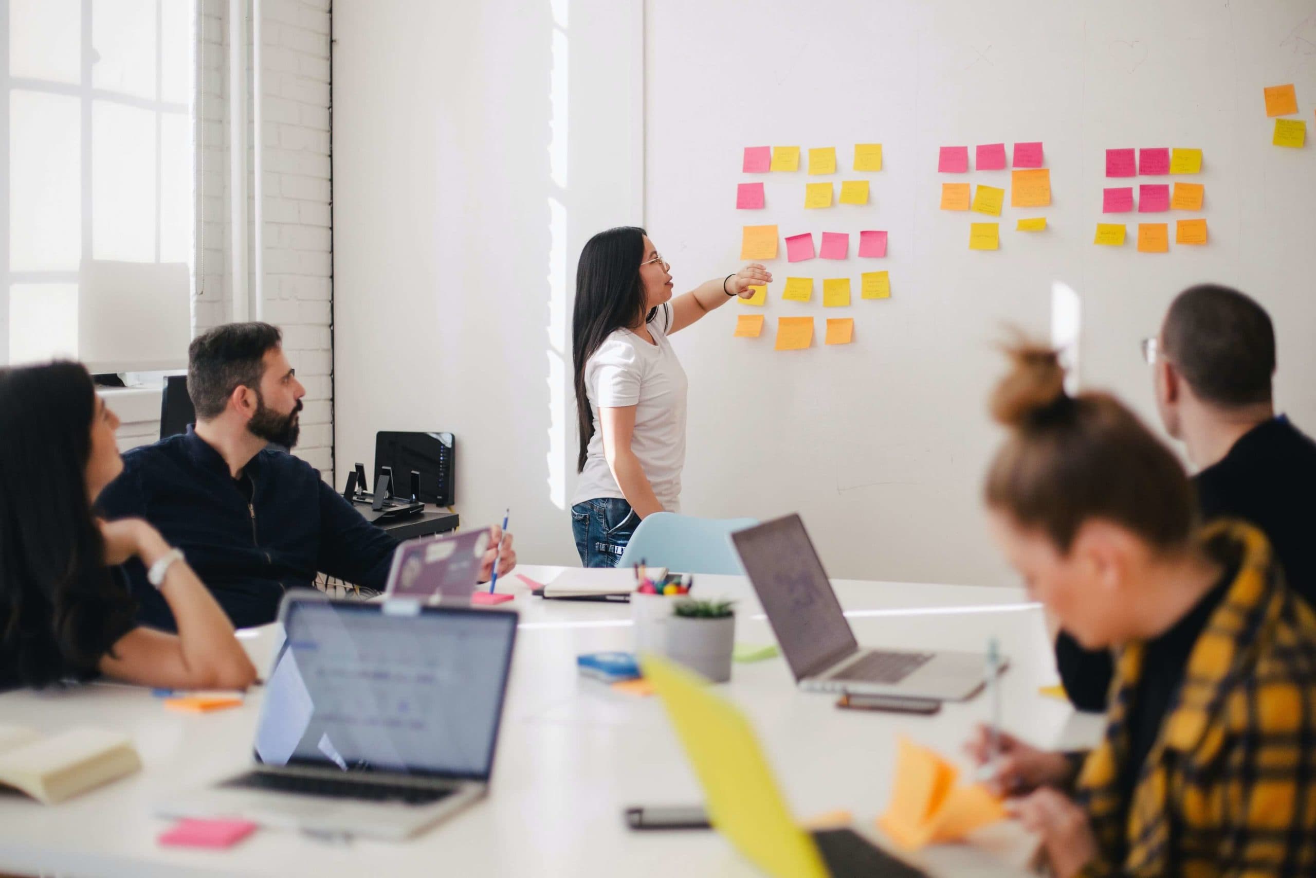 A woman stands in front of a whiteboard covered with colorful sticky notes, presenting to four seated colleagues. The group sits around a table with open laptops and notebooks, engaged in a collaborative meeting in a bright, modern office.