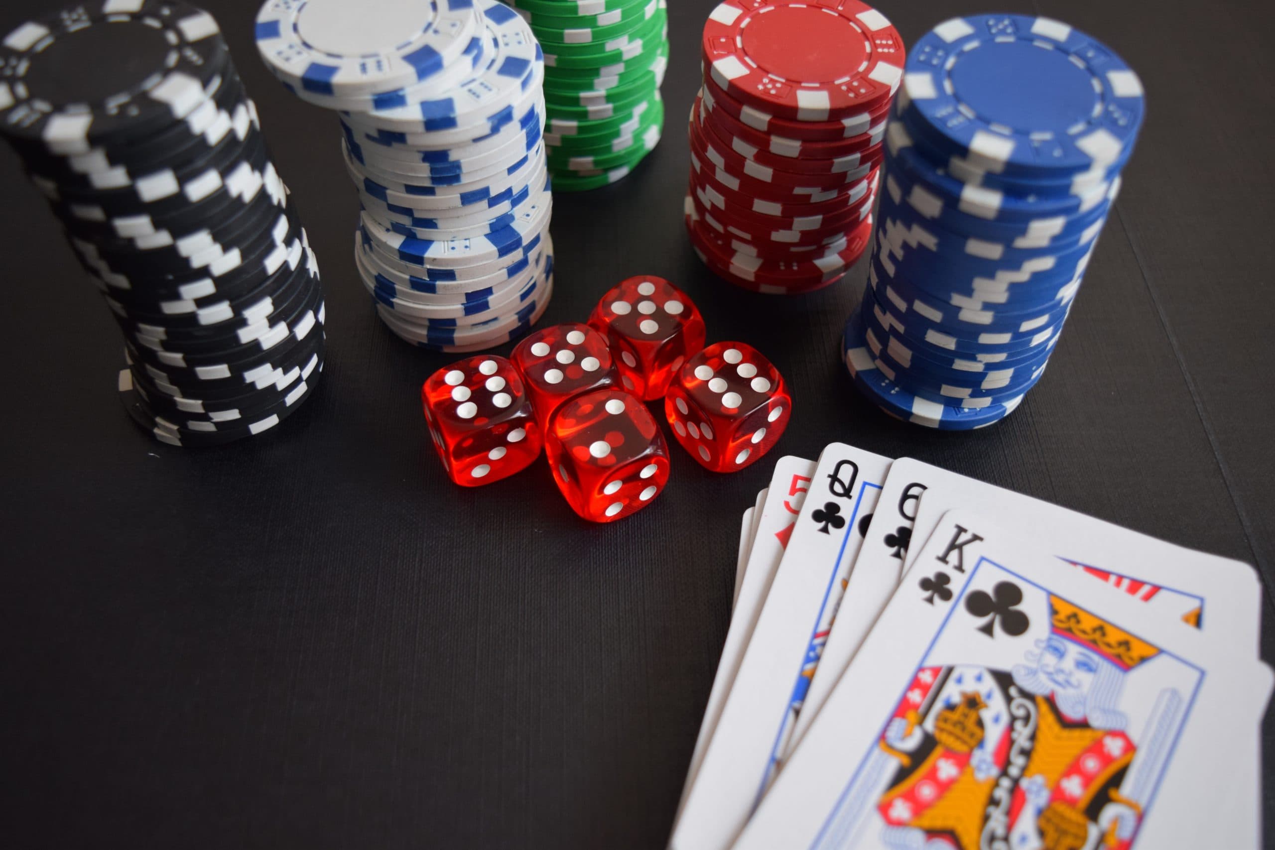 Stacks of poker chips in black, blue, green, and red are arranged on a table alongside red six-sided dice. Nearby, there is a hand of playing cards showing a king, queen, jack, and eight of clubs. The setting suggests a card game.