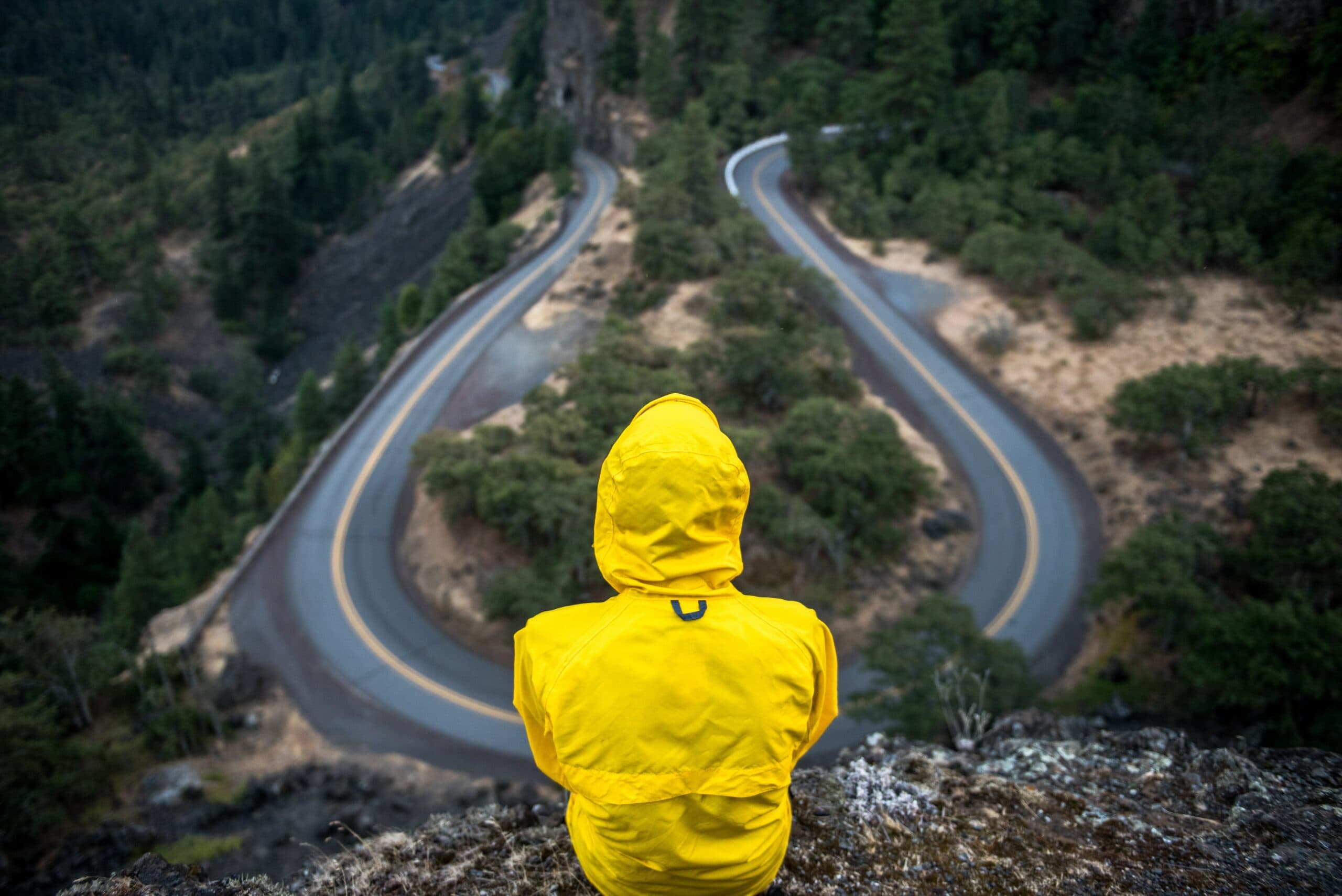 A person wearing a yellow jacket sits on the edge of a cliff, overlooking a winding road surrounded by lush green trees. The scene conveys a sense of adventure and contemplation in a mountainous landscape.