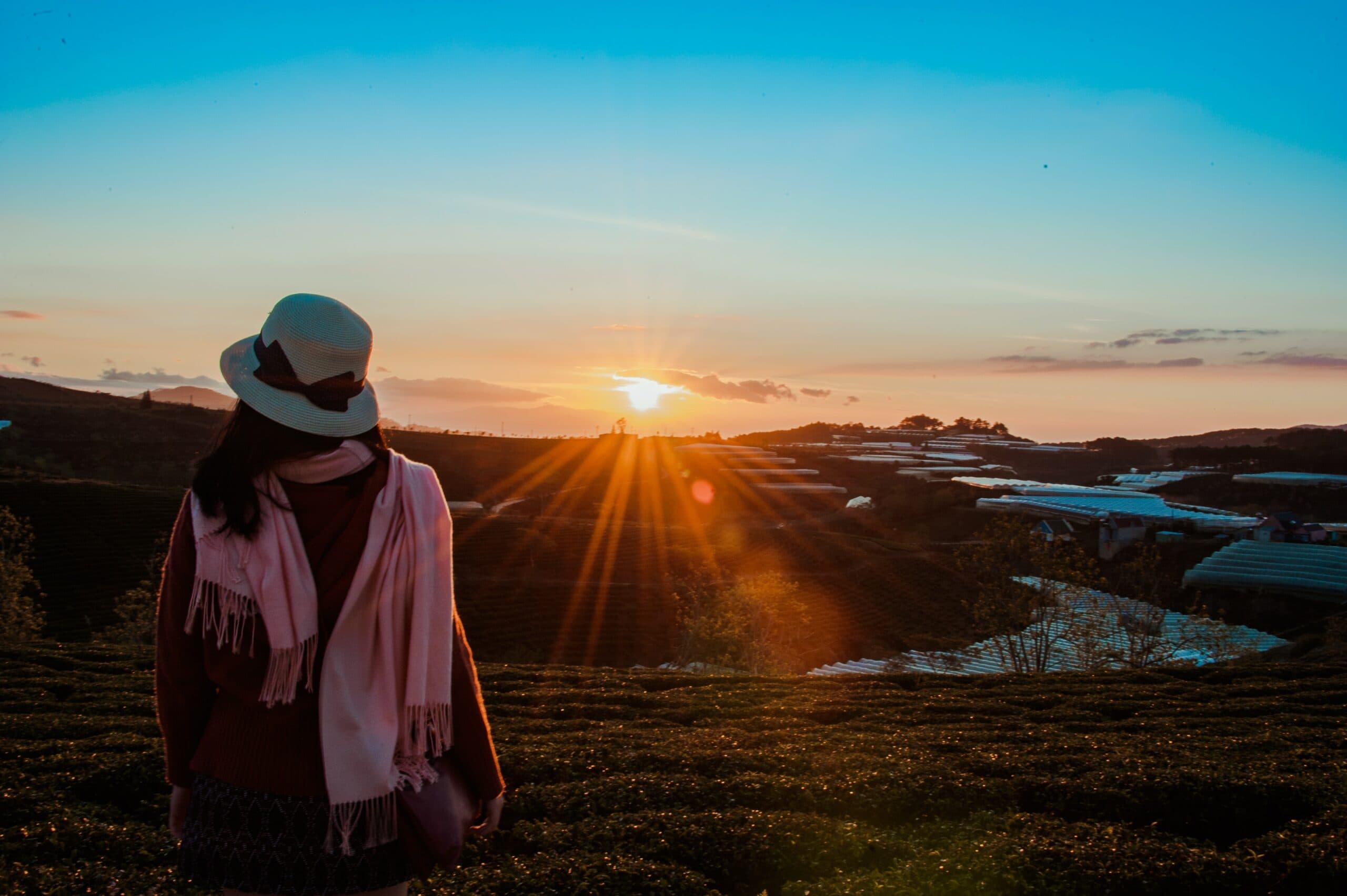 A person wearing a hat and scarf stands in a field, watching a beautiful sunset. The sky is a gradient of blue and orange, with the suns rays spreading outwards. The landscape features rolling hills and reflective surfaces.