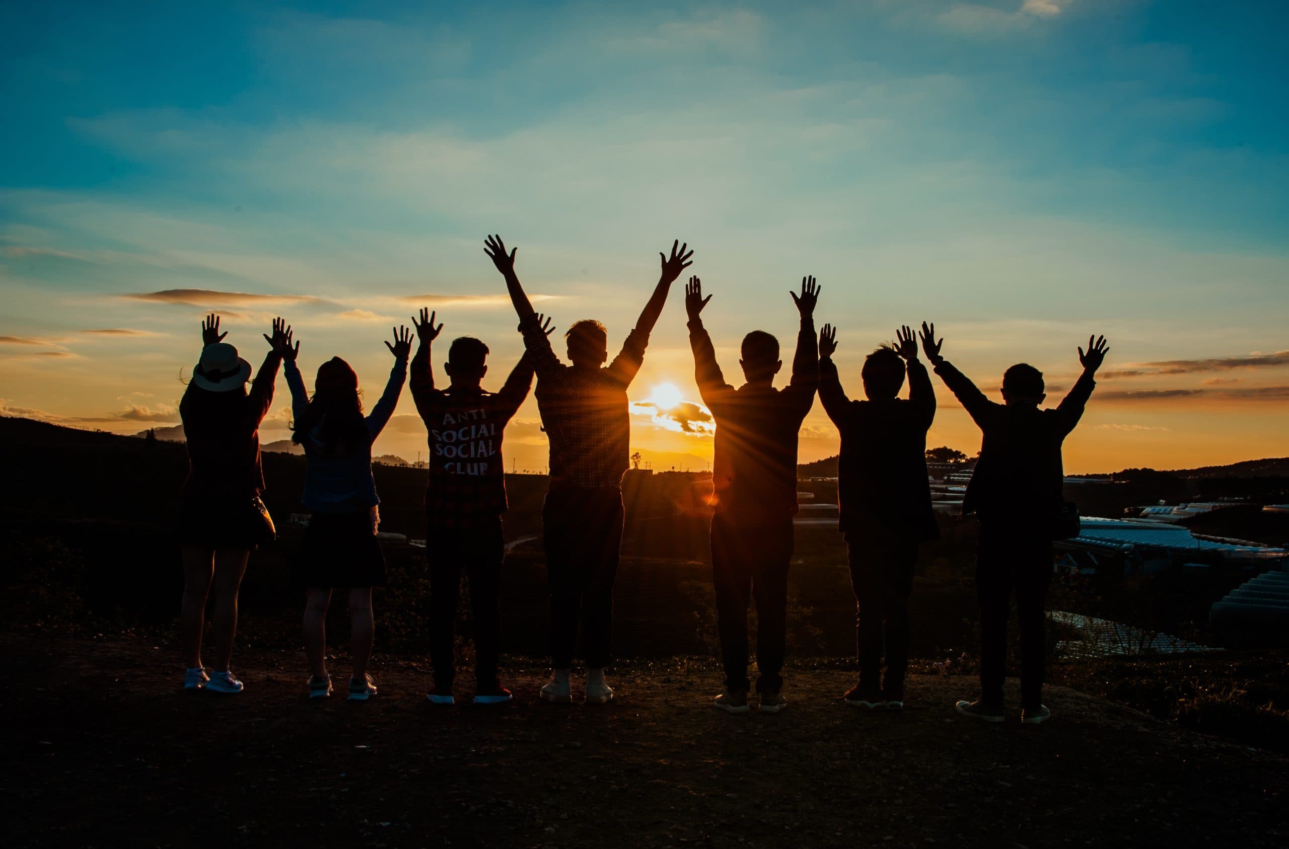 A group of seven people stands on a hill with their backs to the camera, raising their arms in celebration against a sunset sky. The sun sets on the horizon, casting a warm glow and long shadows.