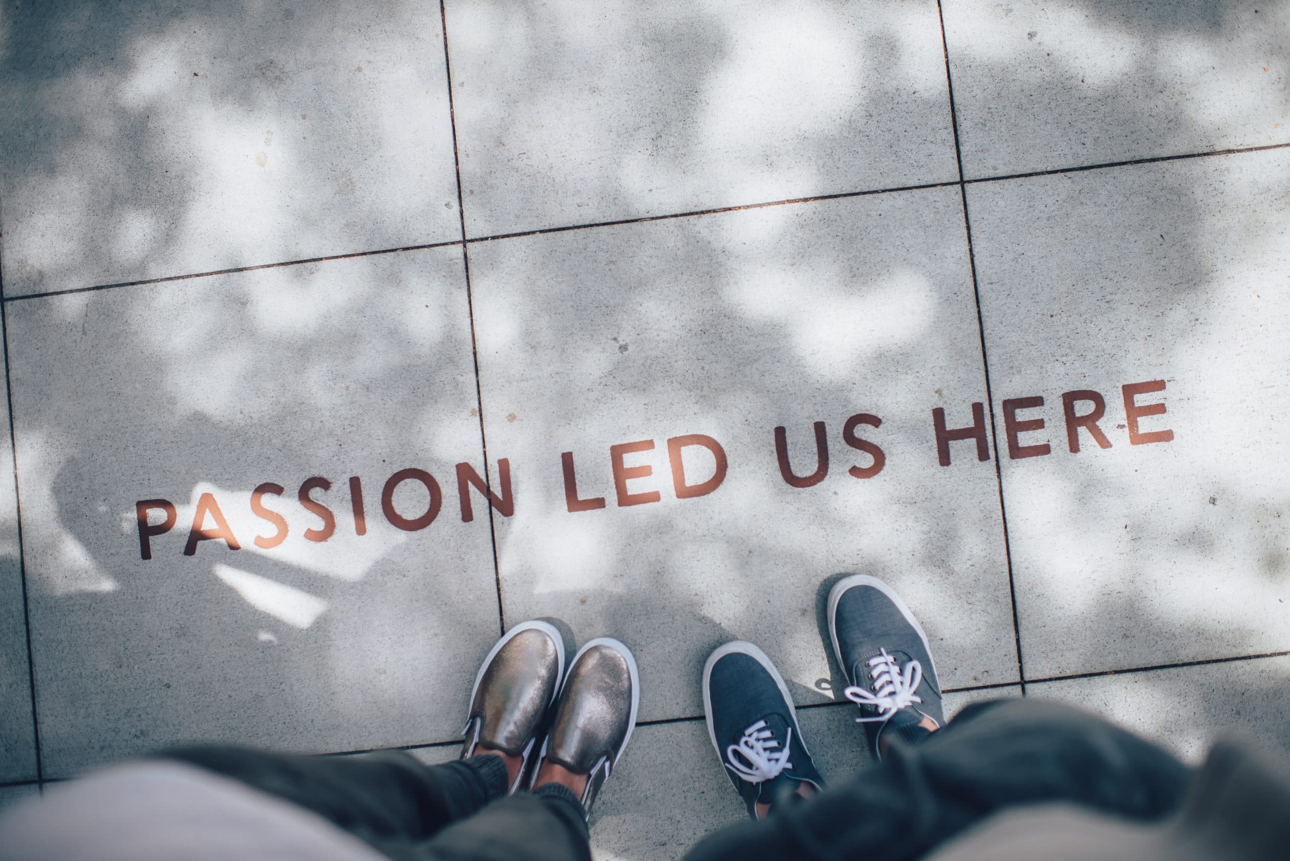 Two pairs of feet stand on a tiled pavement with the text PASSION LED US HERE written in bold letters. Shadows of trees create a dappled effect on the tiles.