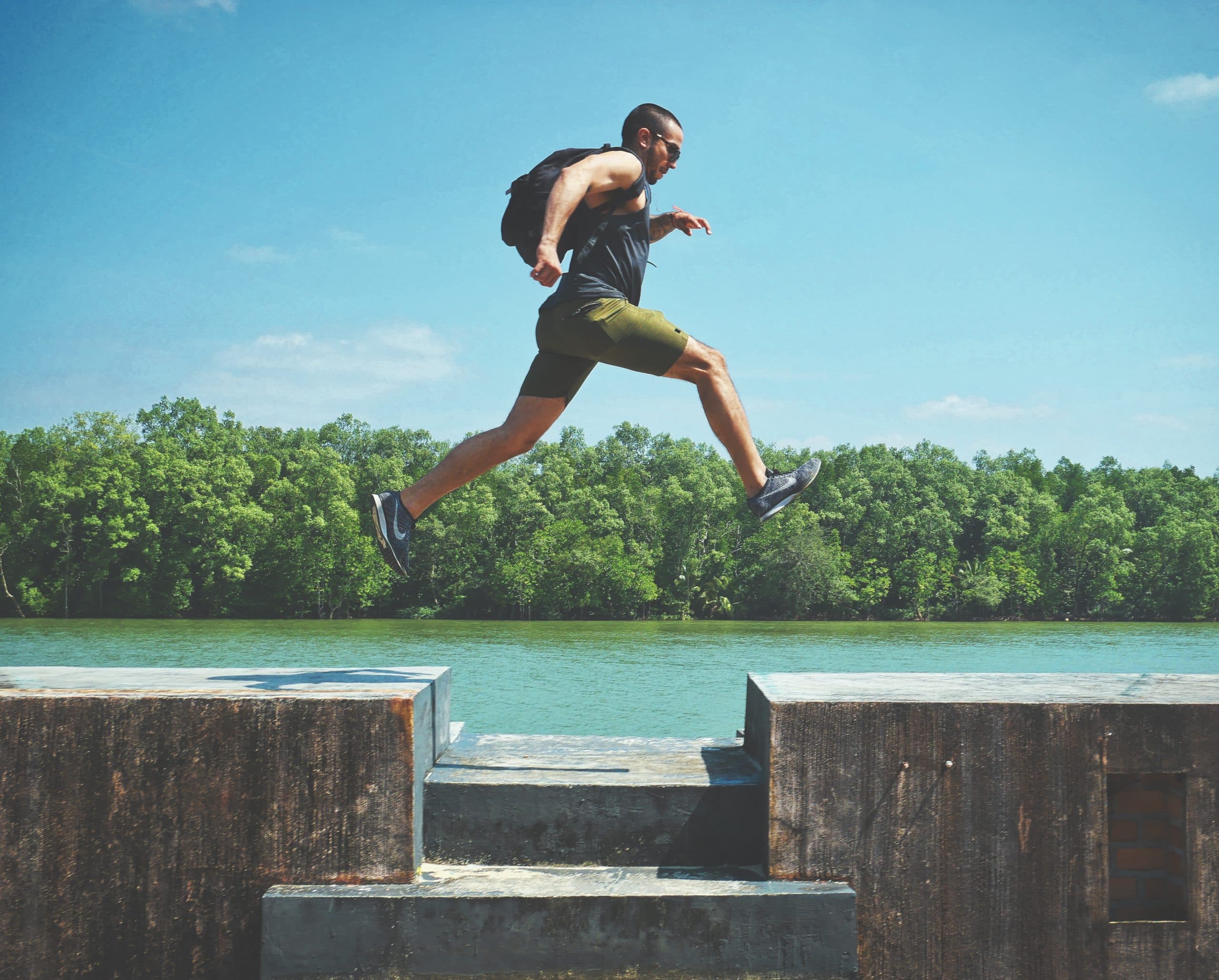 A person wearing a backpack and athletic clothing, mid-air while leaping across a gap between two concrete ledges. A river and lush green trees are visible in the background under a clear blue sky.