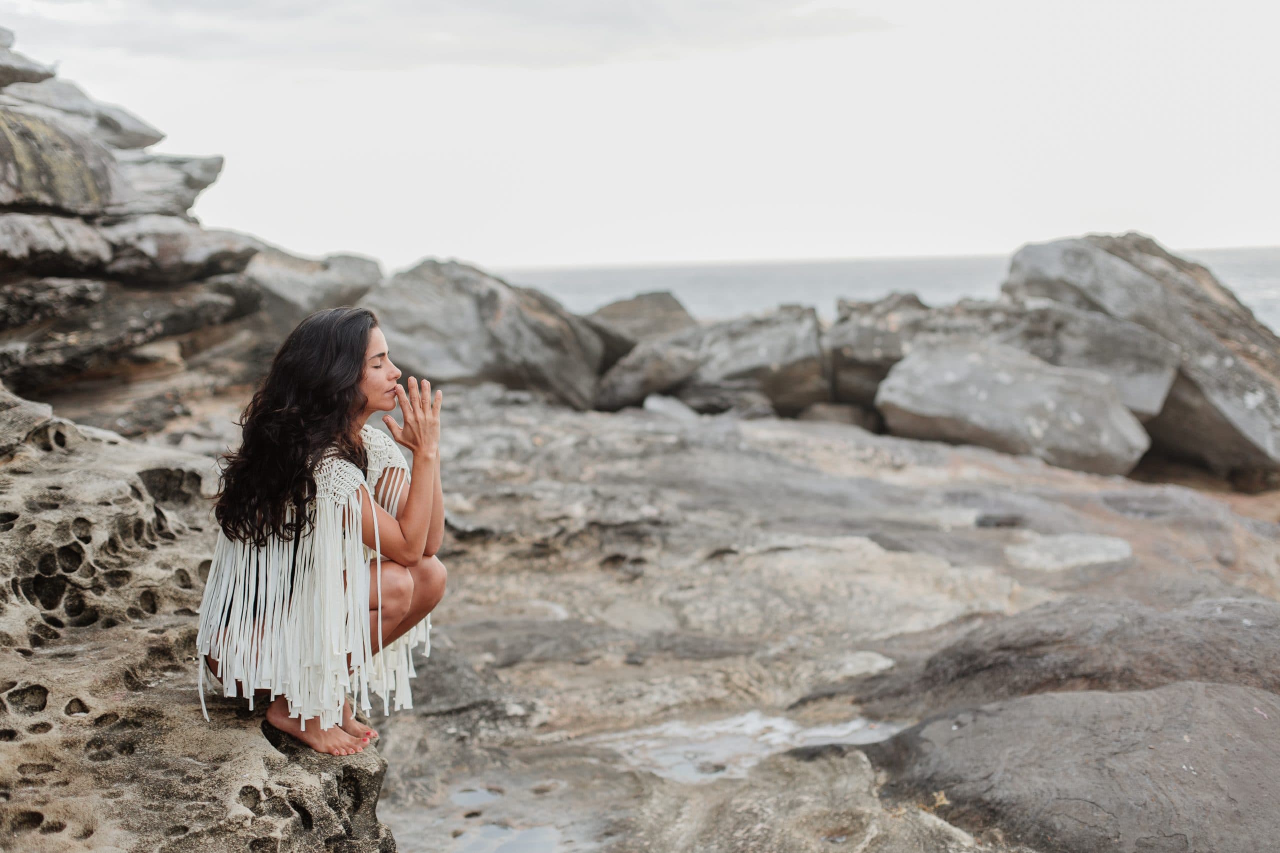 A person with long hair squats on a rocky shoreline, wearing a fringed garment. They have their hands together, gazing toward the ocean under a cloudy sky. The scene conveys a sense of calm and contemplation.