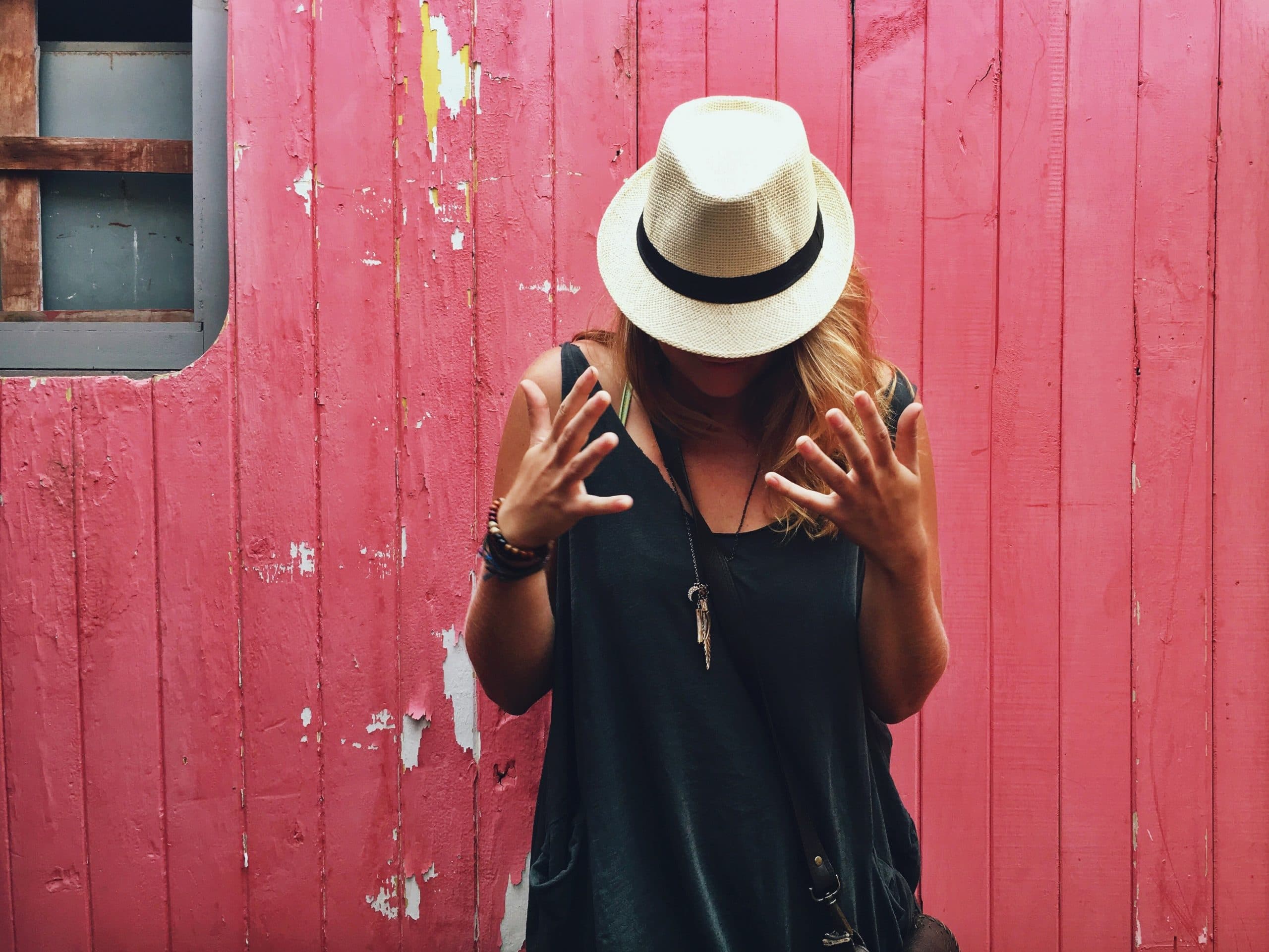 A person wearing a straw hat and dark clothing stands in front of a distressed pink wooden wall. Their face is obscured by the hat, and they are gesturing with both hands spread open.