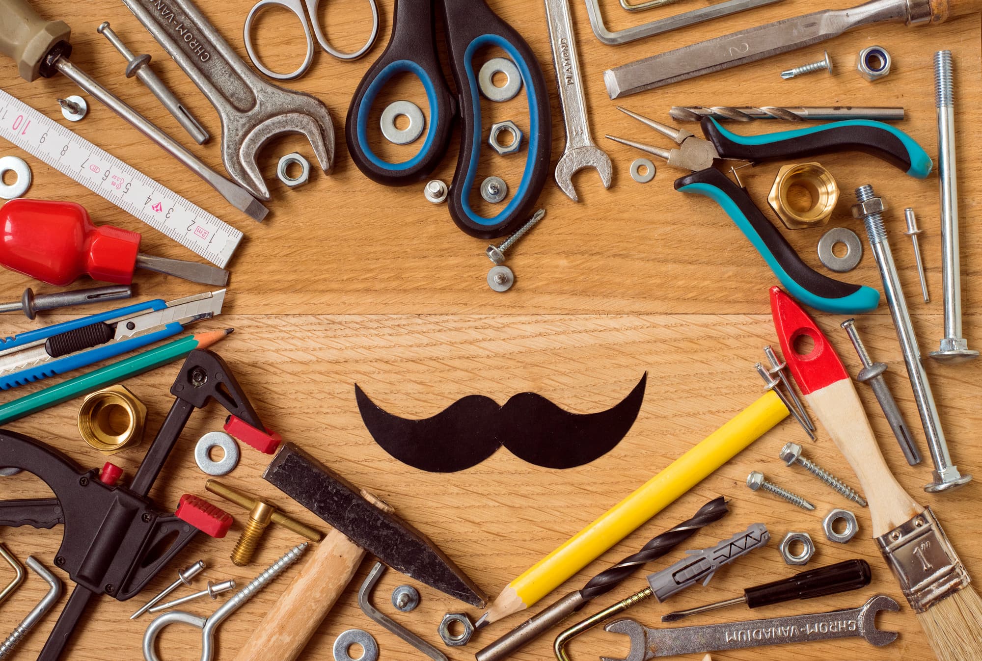 A collection of various tools, including wrenches, pliers, scissors, screws, and a pencil, arranged around a black paper mustache on a wooden surface.