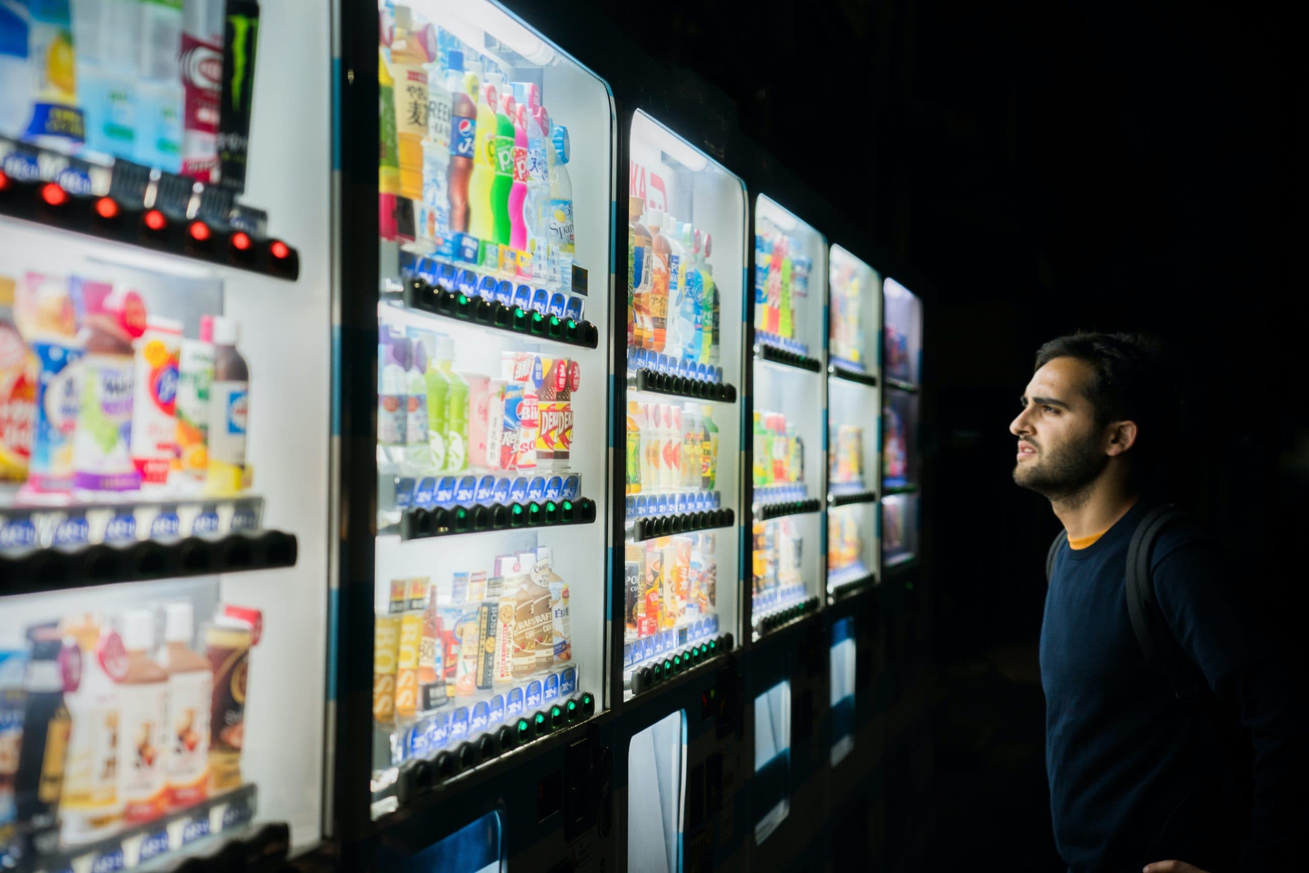 A man stands in front of a brightly lit row of vending machines at night, examining the variety of snacks and drinks on display.