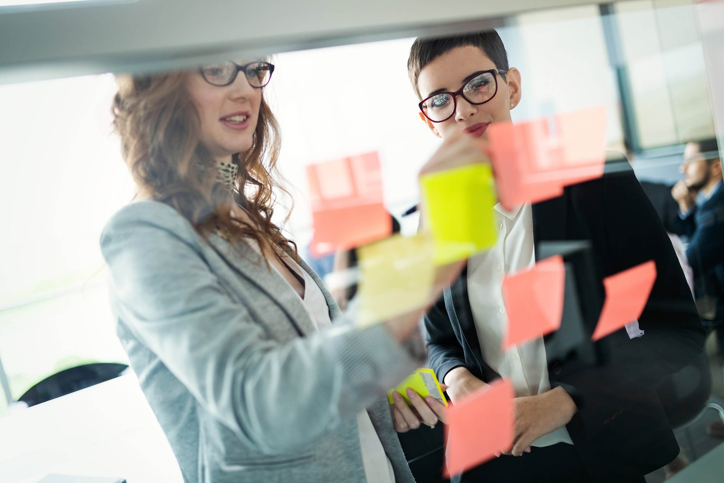 Two people collaborate in an office, placing colorful sticky notes on a glass board. They both wear glasses and business attire, engaging in discussion and planning. The setting is bright and modern.