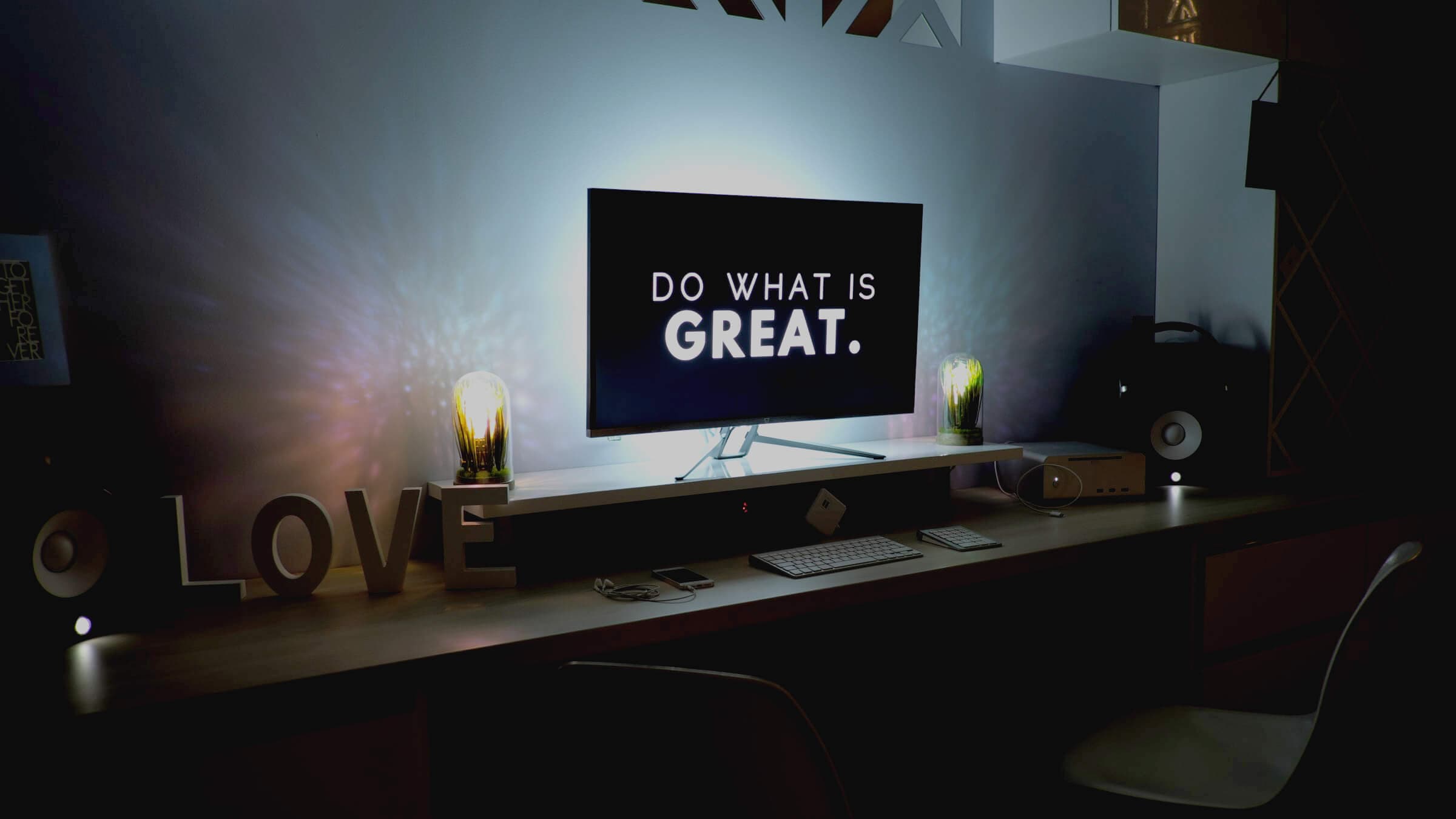 A modern workspace with a computer displaying the text DO WHAT IS GREAT. The desk is decorated with lit bulbs, letters spelling LOVE, and speakers, creating a cozy and motivational atmosphere.