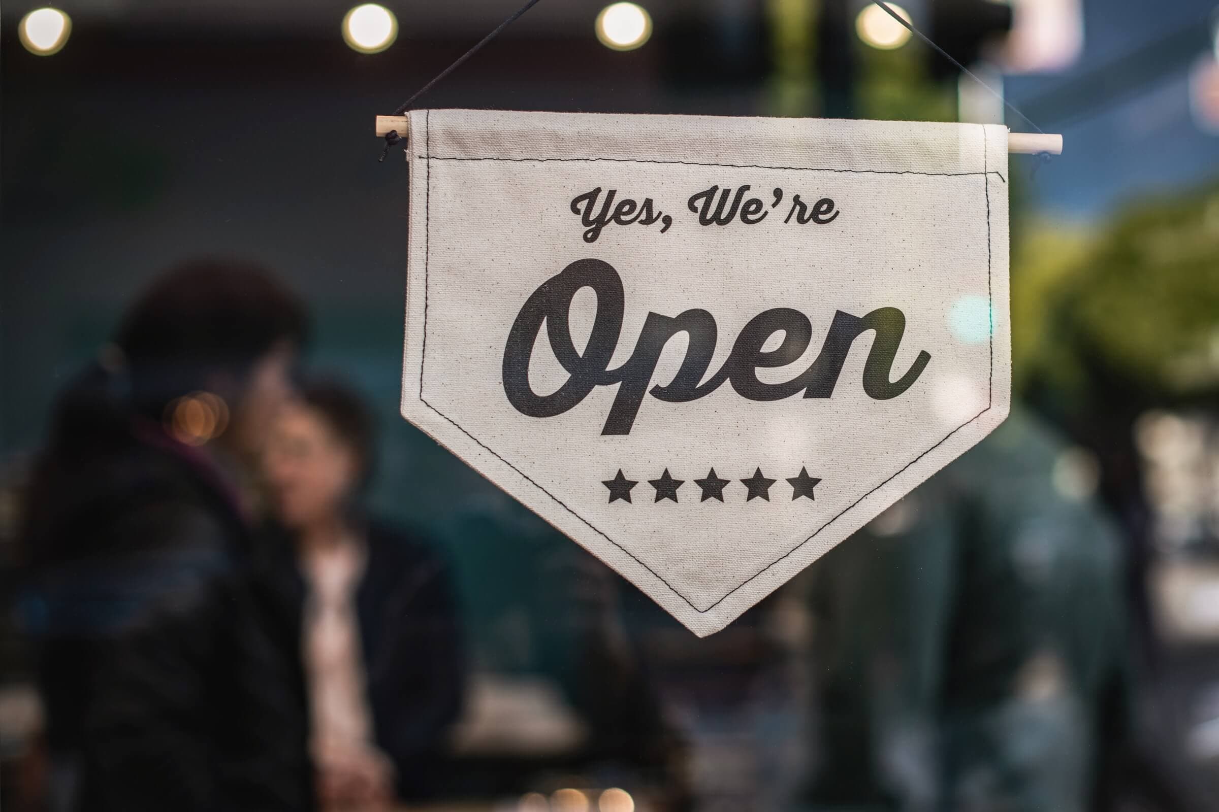 A hanging sign on a glass door reads, Yes, Were Open with five stars below. The background shows a blurred view of people and a tree outside, suggesting a welcoming business environment.