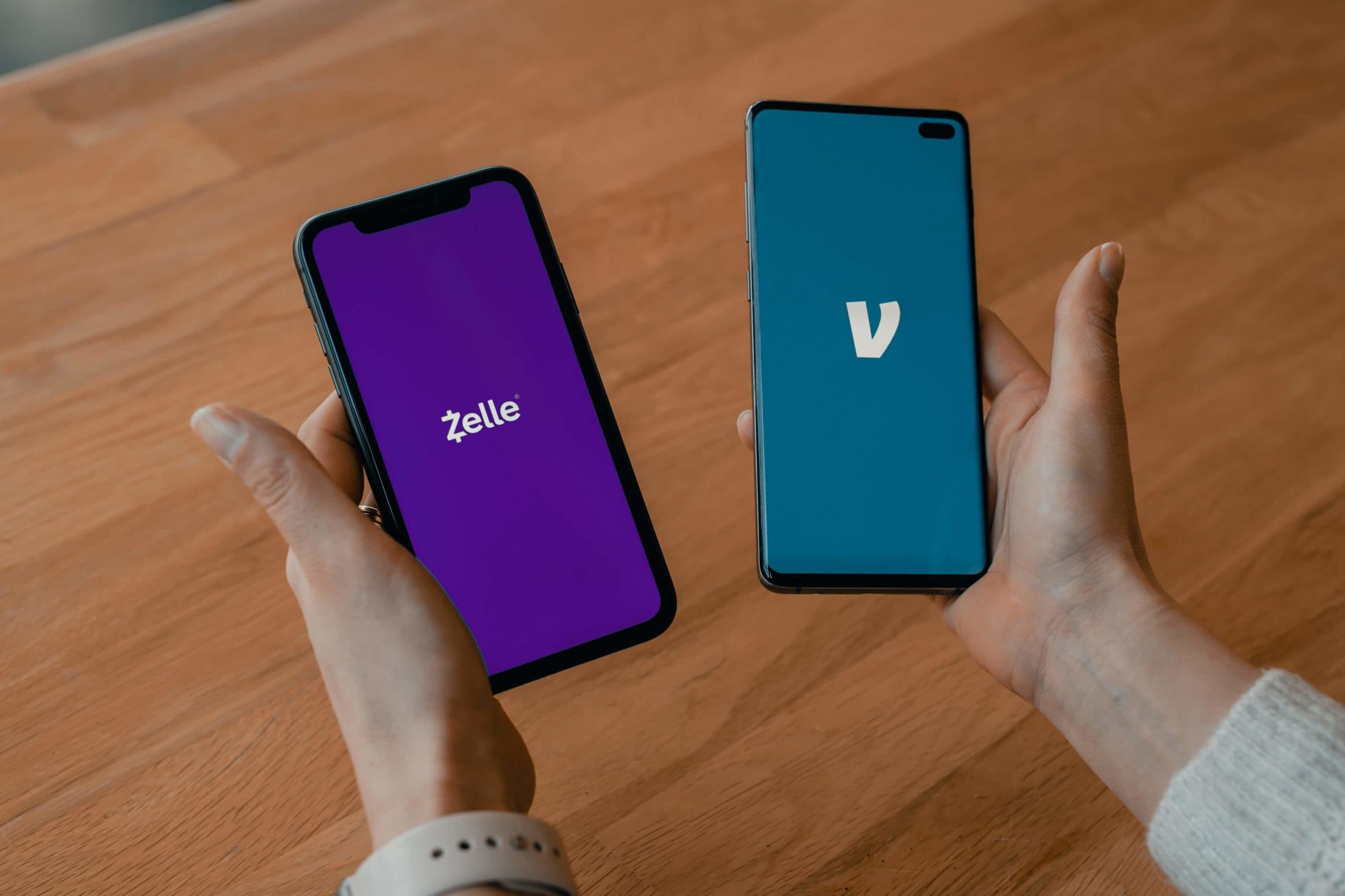 Two hands holding smartphones over a wooden table. The left phone displays the Zelle app with a purple background, while the right phone shows the Venmo app with a teal background. Both hands wear a white wristband.