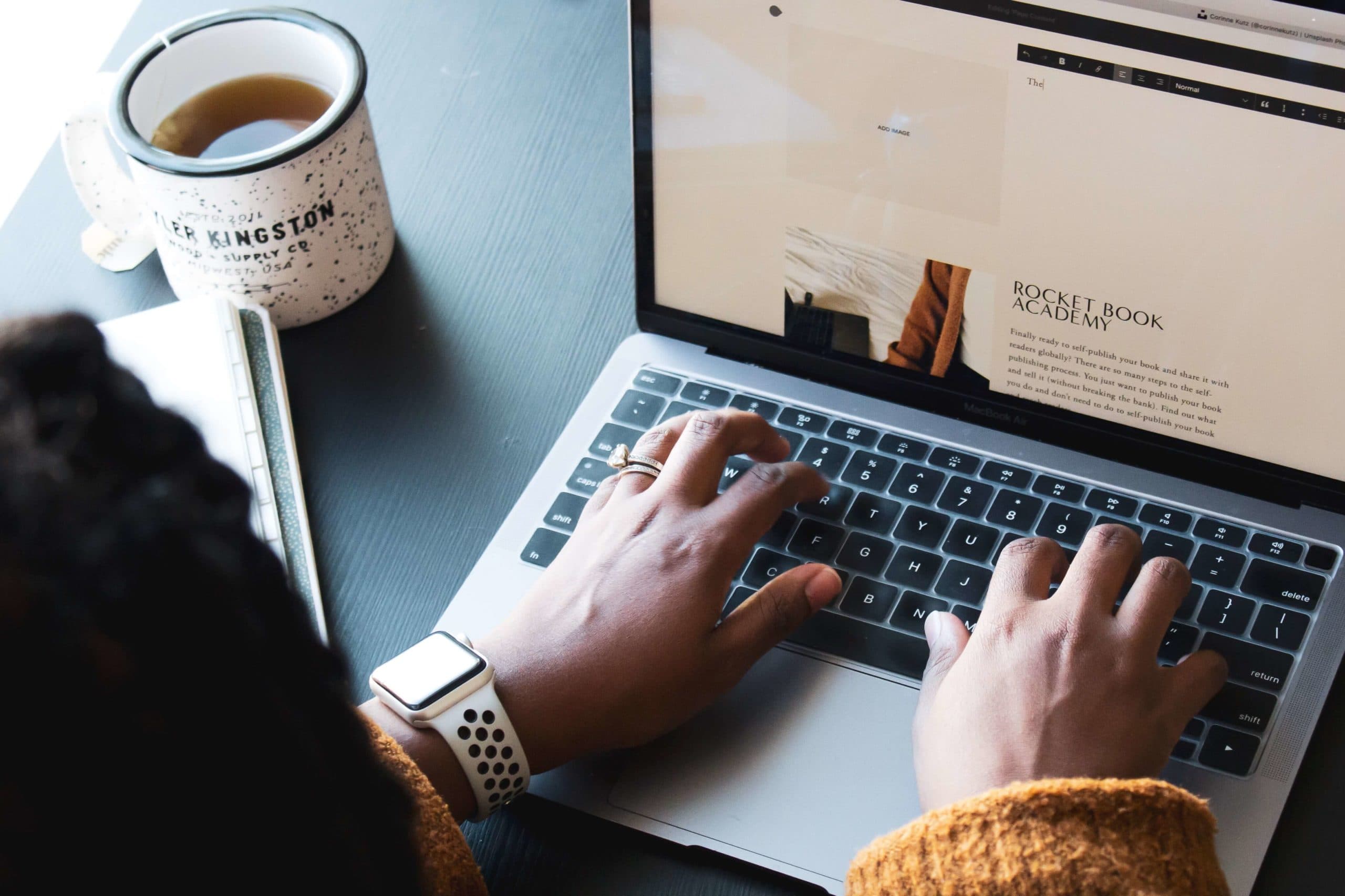 A person wearing a white smartwatch types on a laptop keyboard. A speckled mug filled with a hot beverage sits on the dark table beside them. The screen displays a website titled Rocket Book Academy.