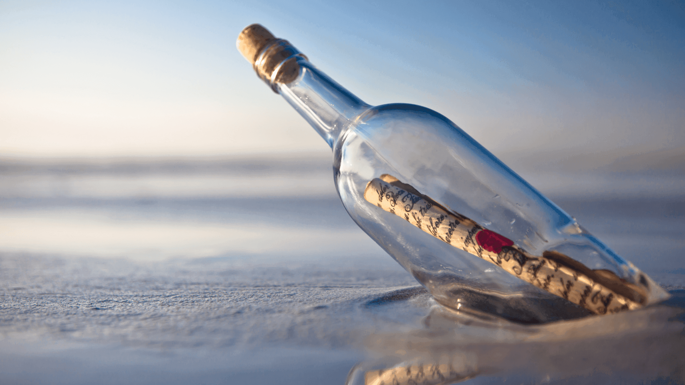 A glass bottle lies on a sandy beach, slightly tilted, with a rolled-up scroll inside. The ocean and sky are visible in the background, creating a serene and hopeful atmosphere.