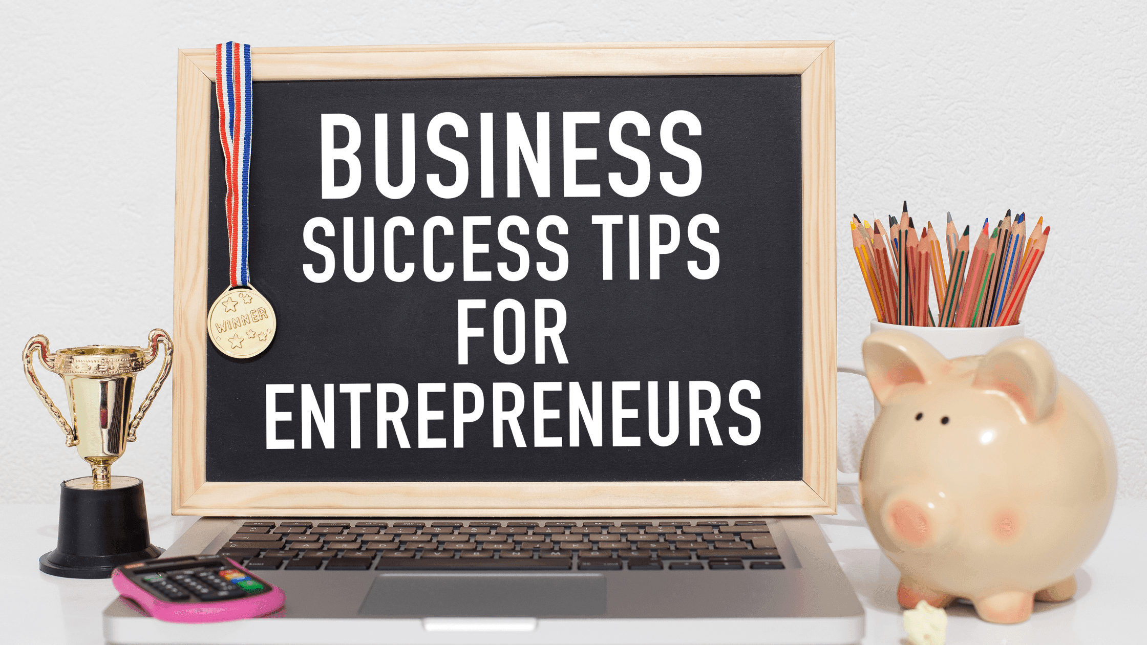 A blackboard on a laptop reads Business Success Tips for Entrepreneurs. A trophy, a medal, a piggy bank, colored pencils, a notepad, and a calculator are placed around the laptop on a desk.