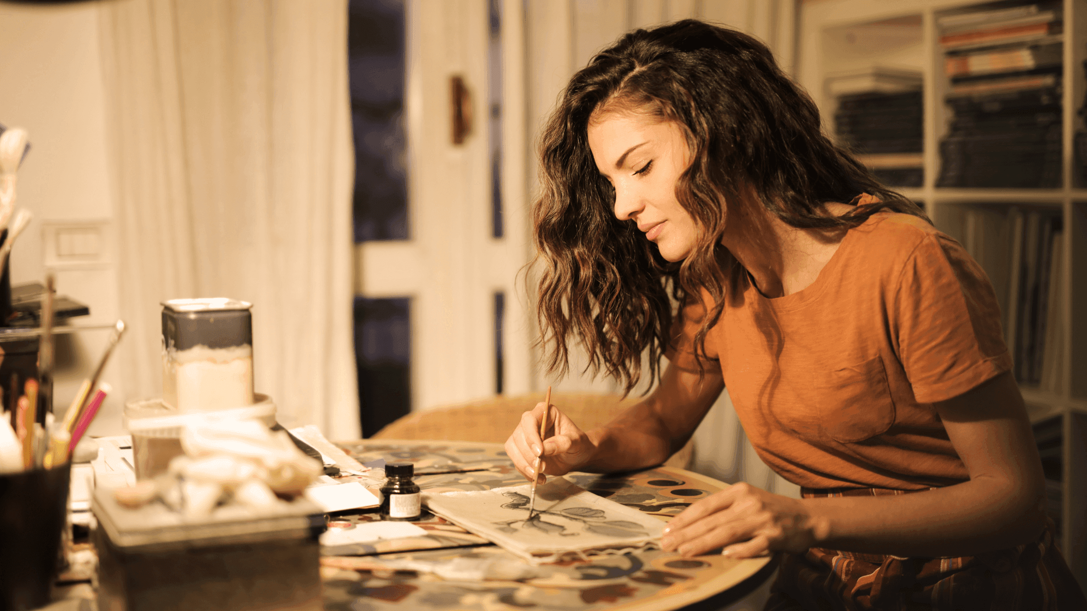 A woman with curly hair sits at a table, focused on painting. She wears a brown shirt and is surrounded by art supplies. The room has bookshelves and a window with curtains in the background.