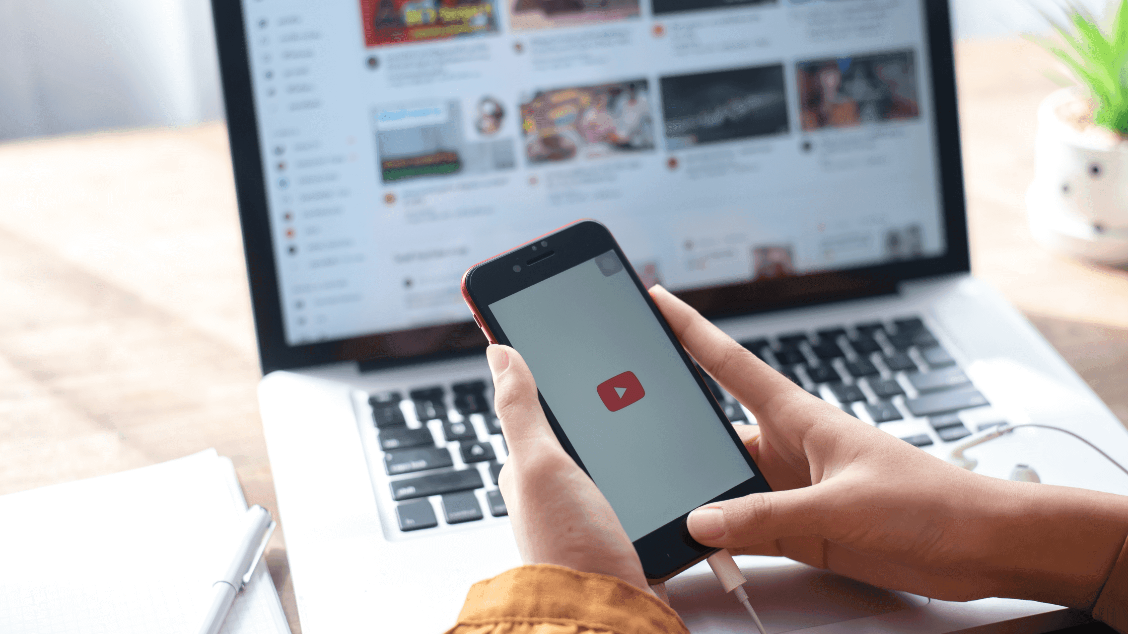 A person holds a smartphone displaying the YouTube logo in front of a laptop showing a YouTube homepage. A notebook and pen are nearby on a wooden table.