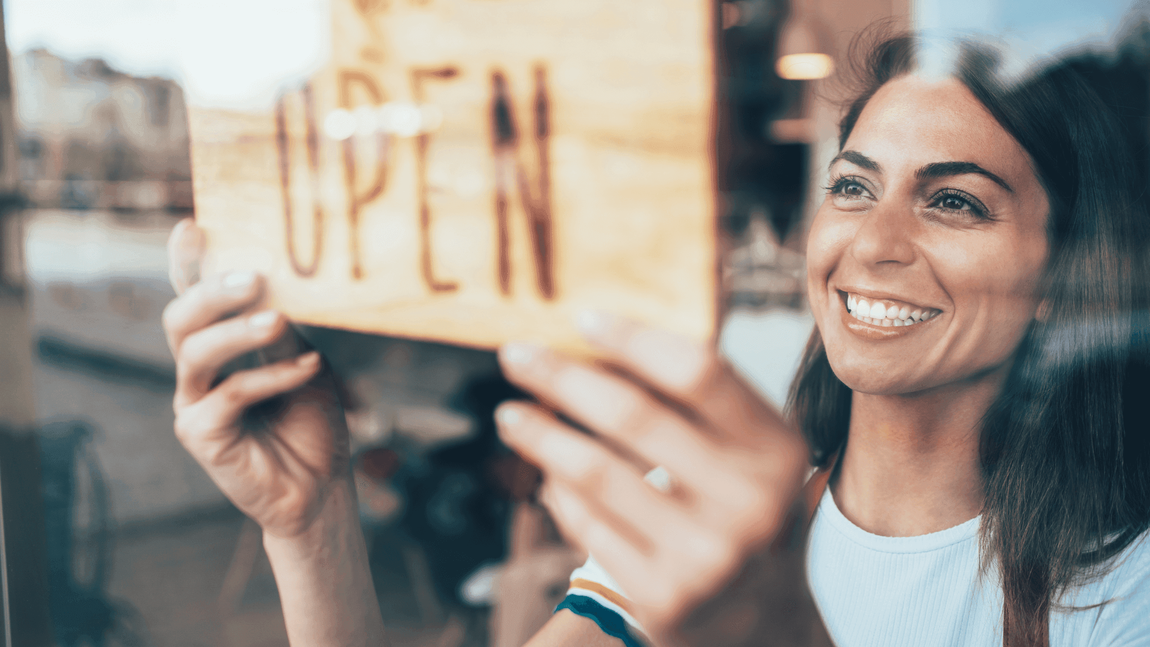 A smiling woman is placing an OPEN sign on a glass door or window. She is wearing a white shirt with colorful stripes on the sleeves. The sign is wooden, and the background shows an outdoor scene slightly out of focus.