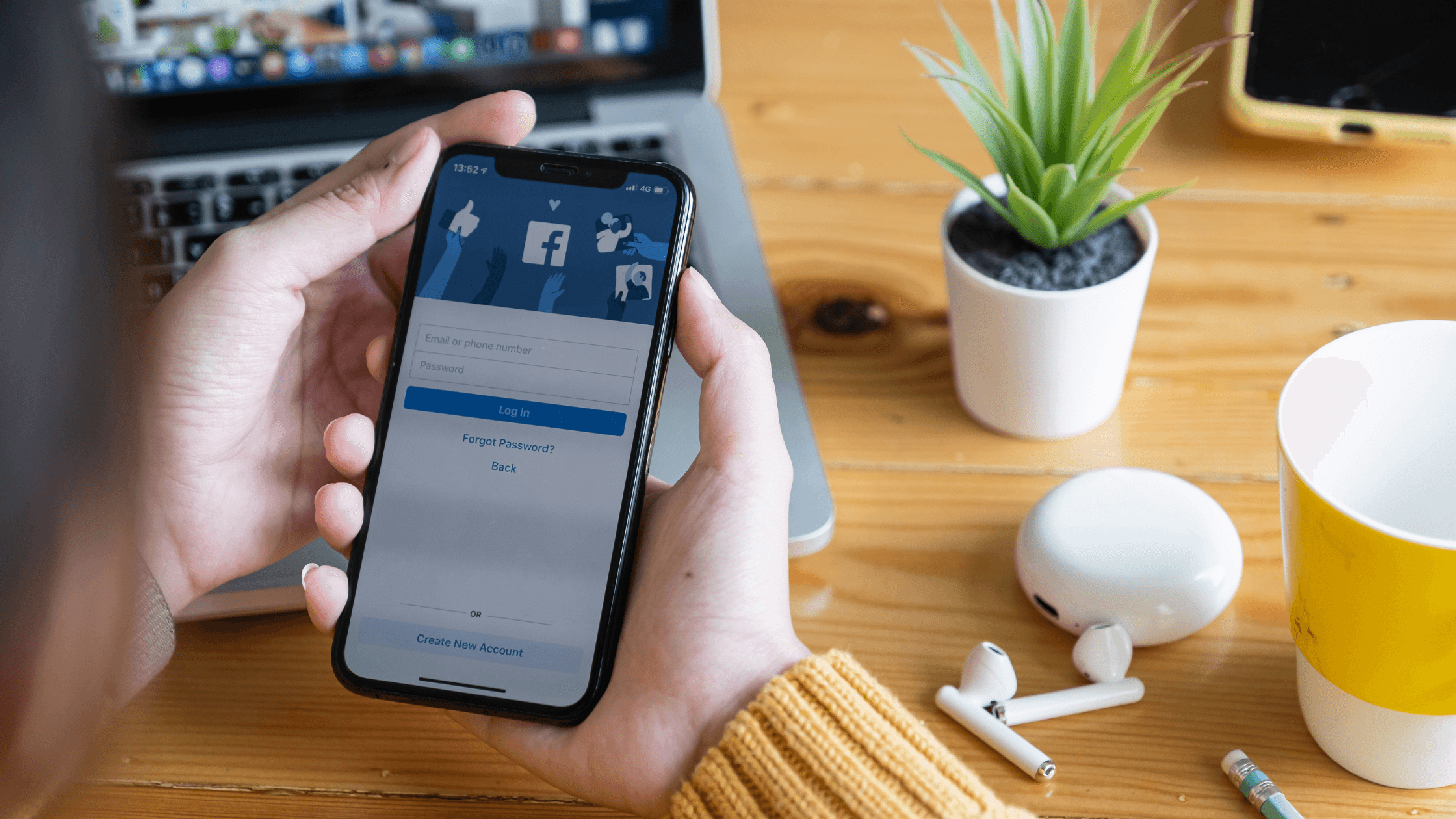 A person holds a smartphone displaying a Facebook login screen. On the wooden table are a laptop, a small potted plant, a pair of white wireless earbuds, and a yellow mug.