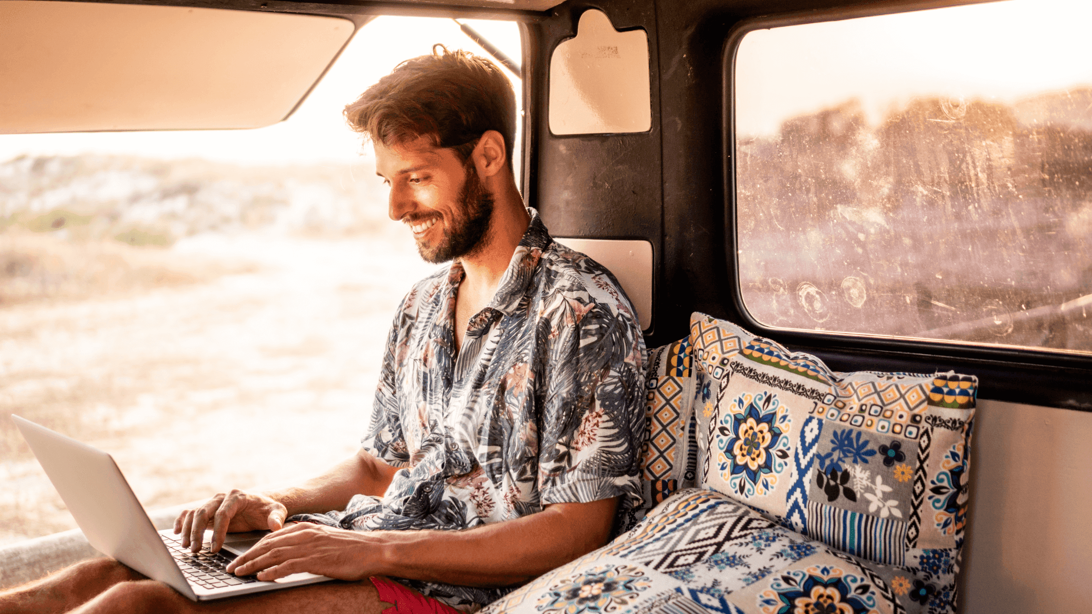 A man sits inside a camper van, smiling while using a laptop. The interior is cozy with patterned cushions and sunlight streams through the window, illuminating the scenic landscape outside.