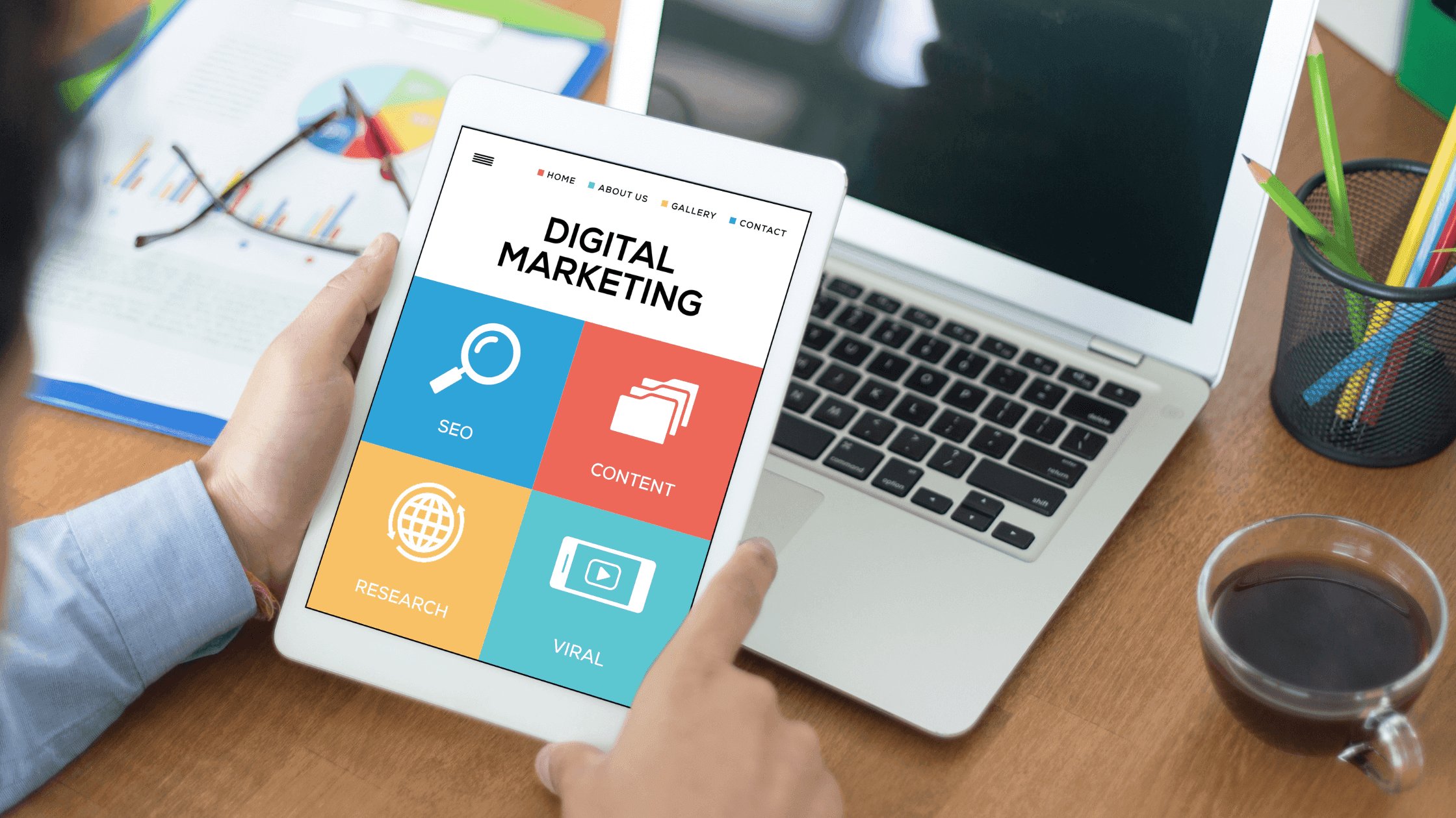 A person holding a tablet displaying a digital marketing app with icons for SEO, content, research, and viral strategies. A laptop, colorful charts, pens, and a cup of coffee are on the wooden desk. The setting is a modern workspace.