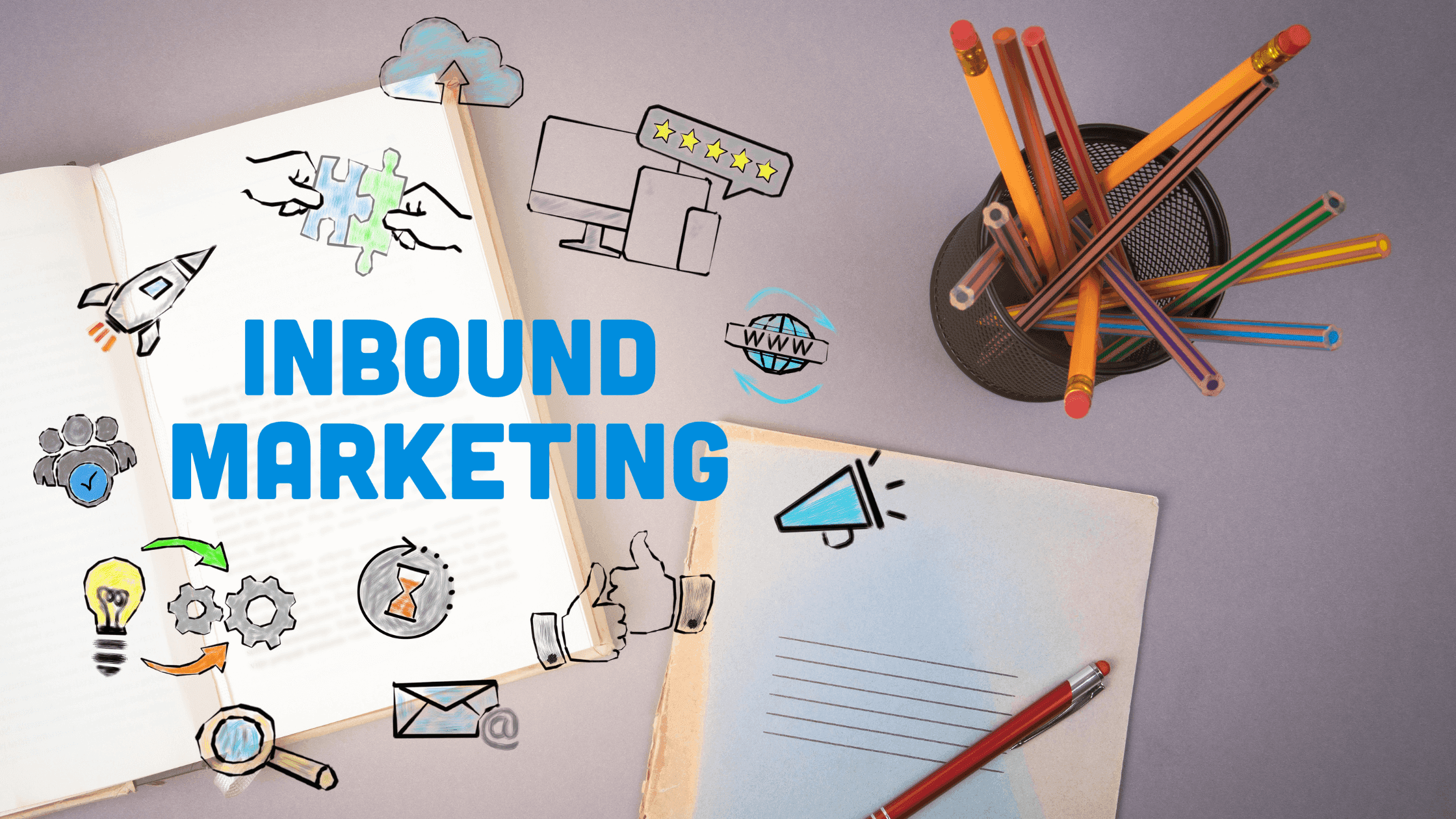 An open notebook with digital marketing icons such as a rocket, gears, and an envelope surrounding the text Inbound Marketing. Nearby, a closed notebook and a container holding colorful pencils are on a gray surface.