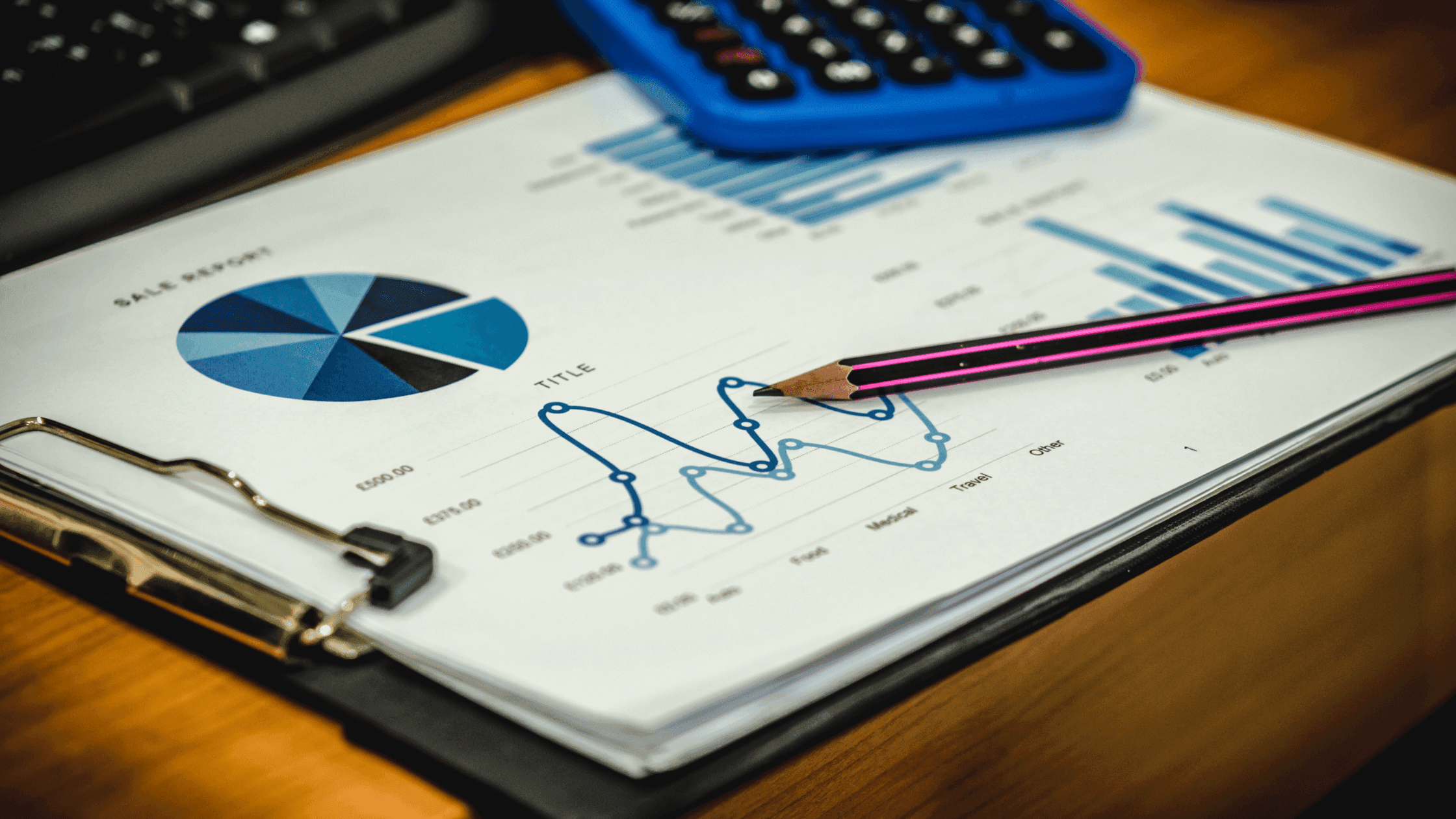 A clipboard holds a sales report with charts and graphs, including a pie chart and a line graph. A pencil rests on the paper, and a calculator is partially visible on a wooden desk.