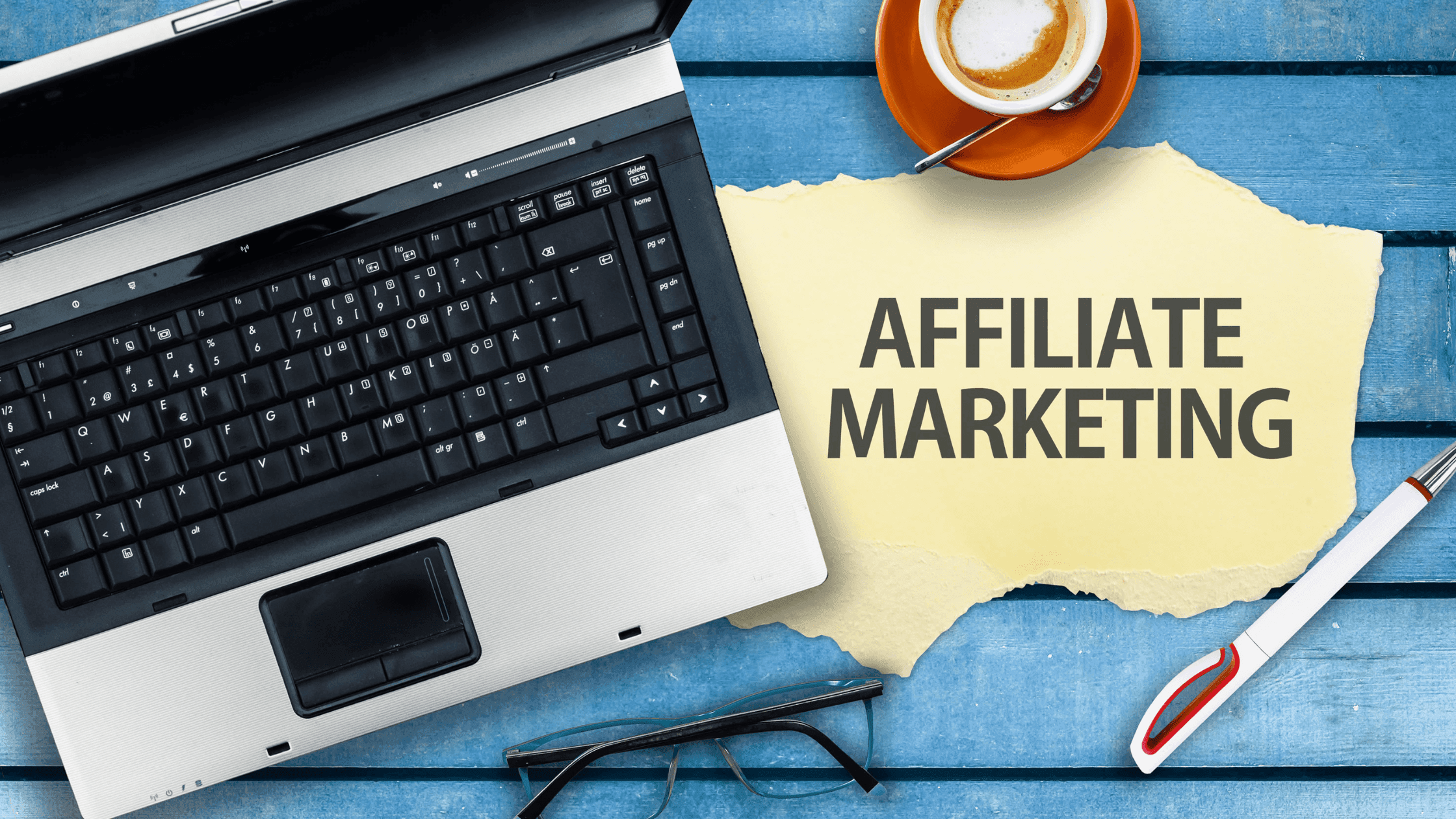 A laptop rests on a blue wooden table next to a cup of coffee. A yellow paper reads Affiliate Marketing. Nearby are glasses and a pen.