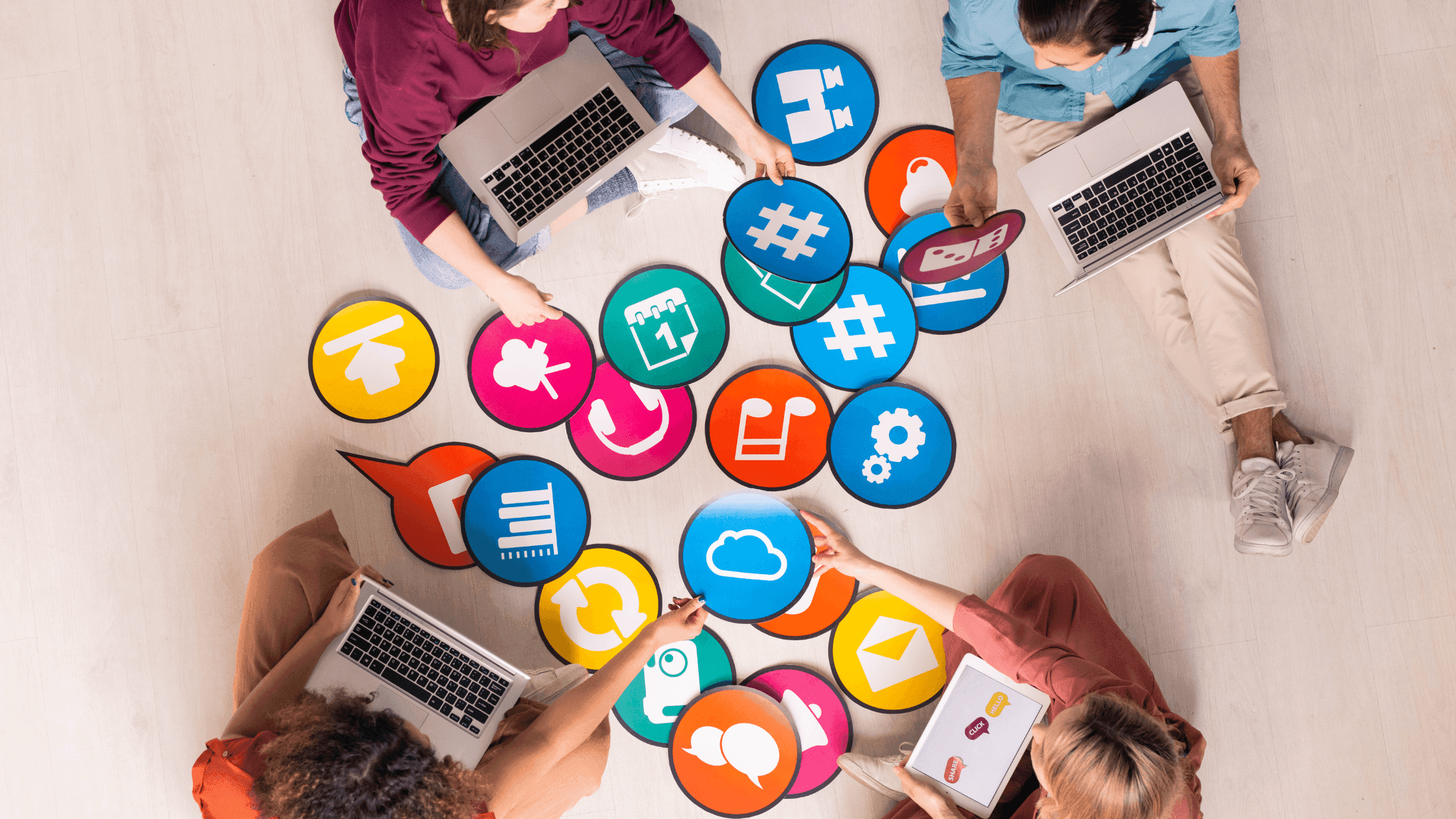 Four people sit on the floor with laptops, surrounded by colorful icons representing various social media platforms and digital symbols, such as hashtags, clouds, and notifications.