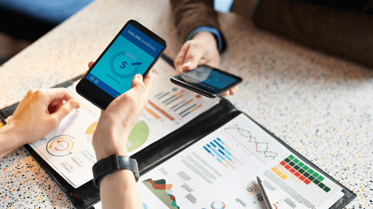 Two people sitting at a table, each holding a smartphone displaying graphs. There are charts and graphs on paper in a binder on the table. The setting suggests a discussion or meeting about financial data or business analytics.