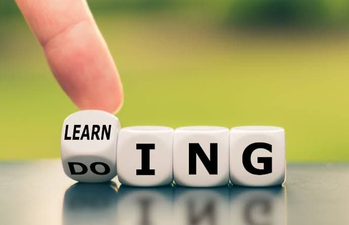 A hand flips a cube changing the word DOING to LEARNING, symbolizing the phrase learning by doing. The background is blurred green.