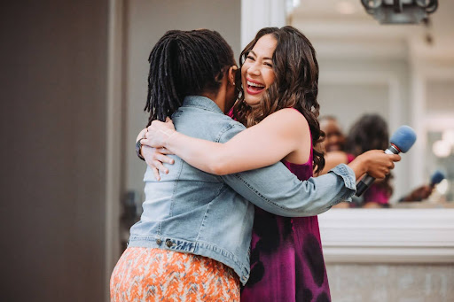 Two women embrace warmly. One woman has her back to the camera and wears a denim jacket; the other, in a purple dress, smiles joyfully and holds a blue microphone. A mirror behind them reflects their hug.