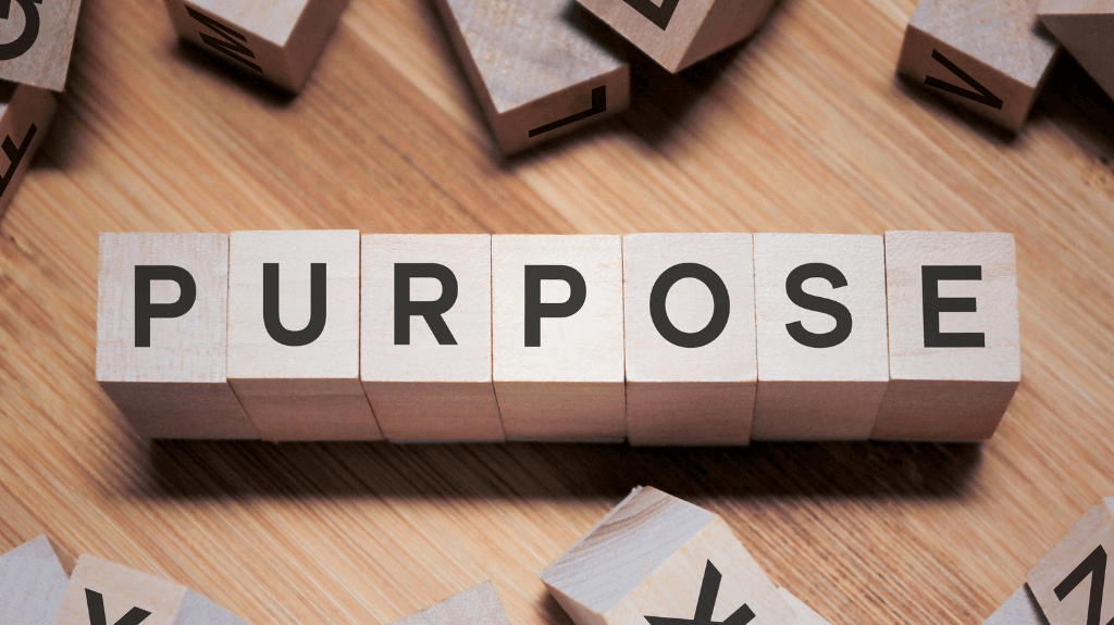Wooden blocks spell out the word PURPOSE on a wooden surface, surrounded by scattered blocks with letters.