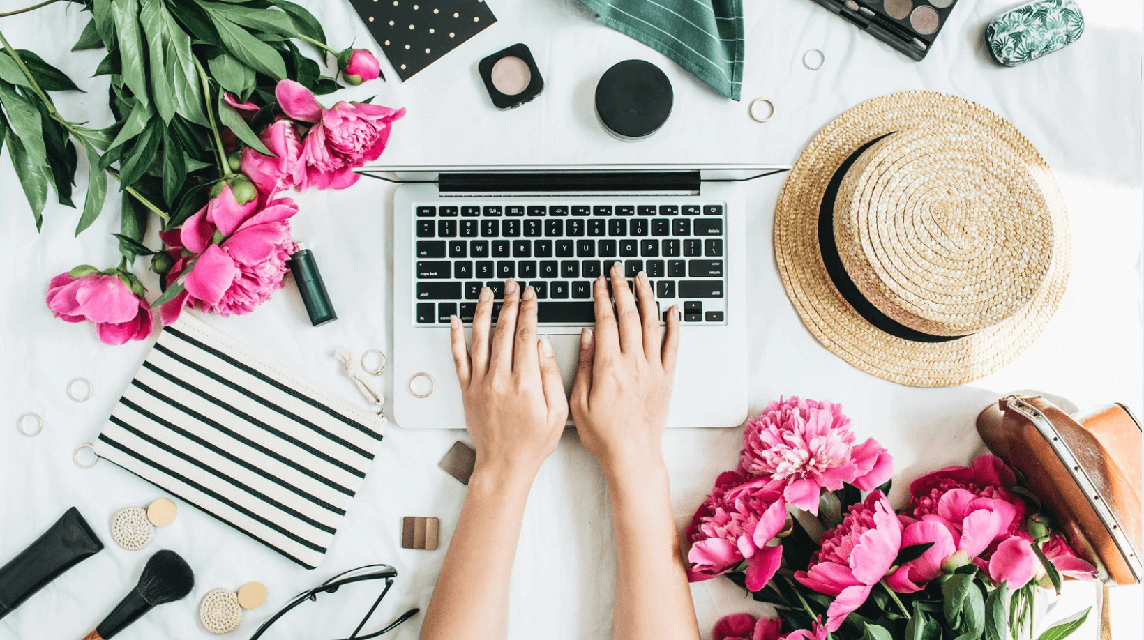 Hands typing on a laptop surrounded by pink flowers, a straw hat, makeup items, a striped pouch, sunglasses, and a leafy plant on a white surface.