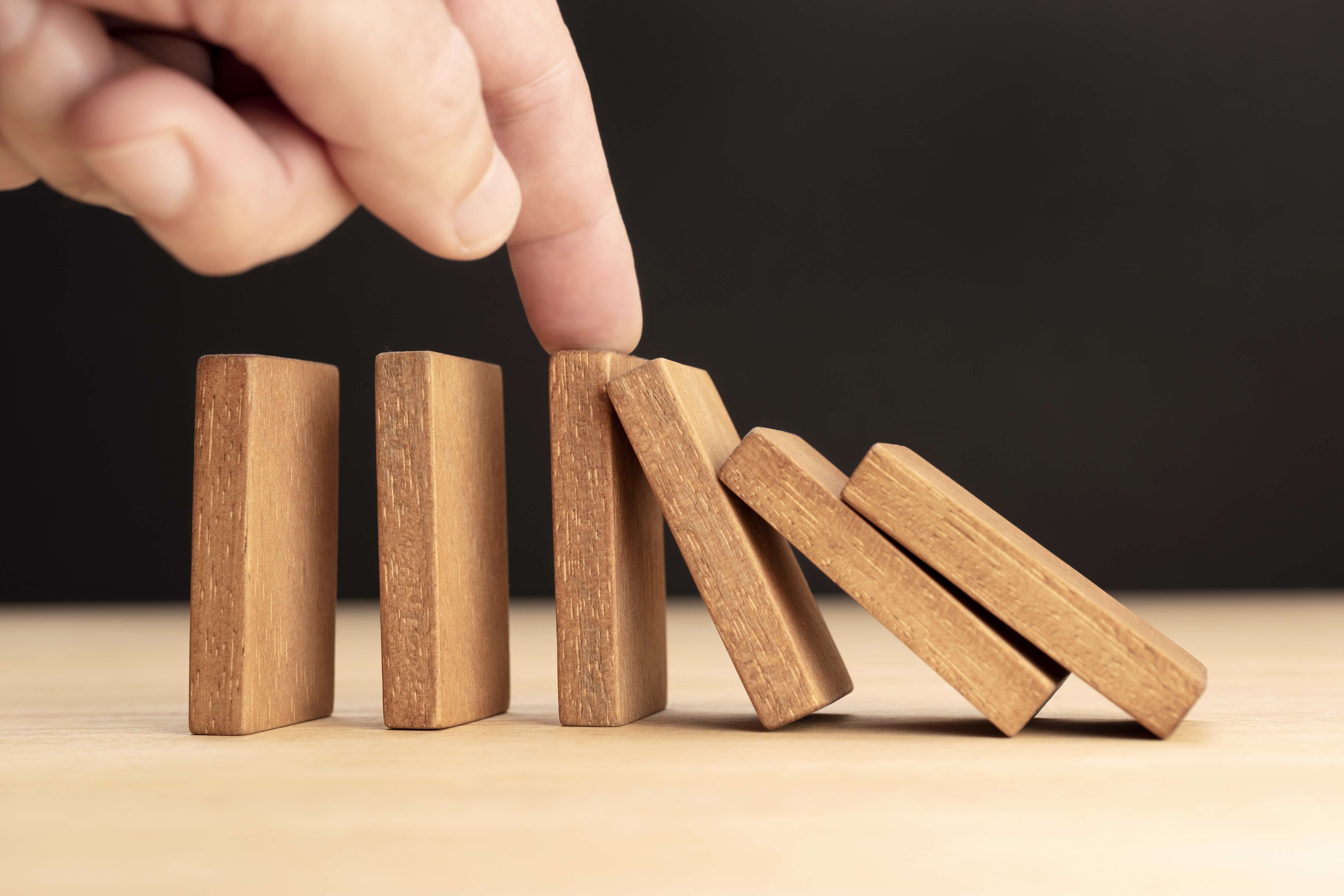 A hand stops a row of falling wooden dominoes, preventing the rest from toppling over on a light-colored surface with a dark background.