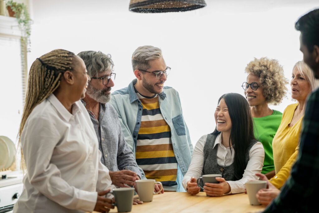 A diverse group of adults stand around a kitchen table, smiling and laughing together while holding coffee mugs in a bright, cozy setting.